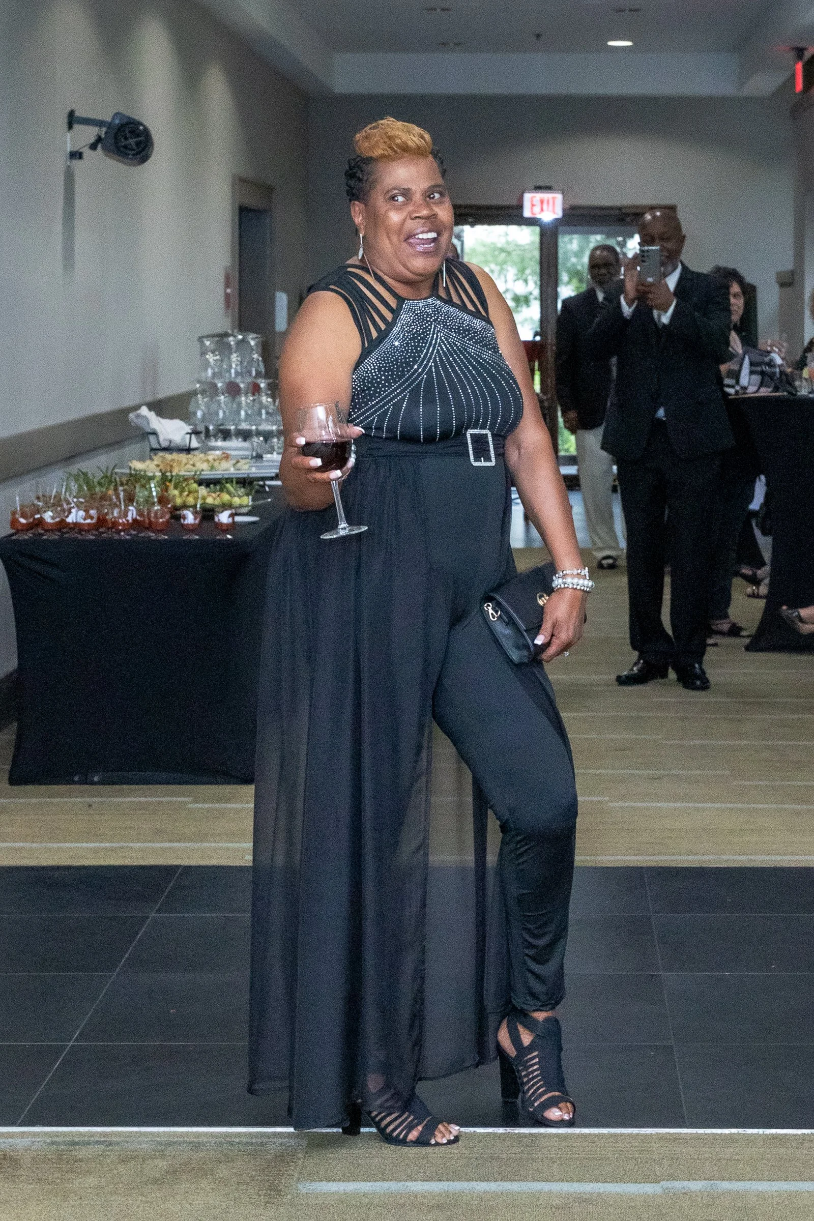 A woman in a black dress with sheer details and a belt, holding a glass of red wine, standing in a banquet hall with a table of appetizers behind her. She has short blonde and black hair and is smiling. There are several people in formal attire, some