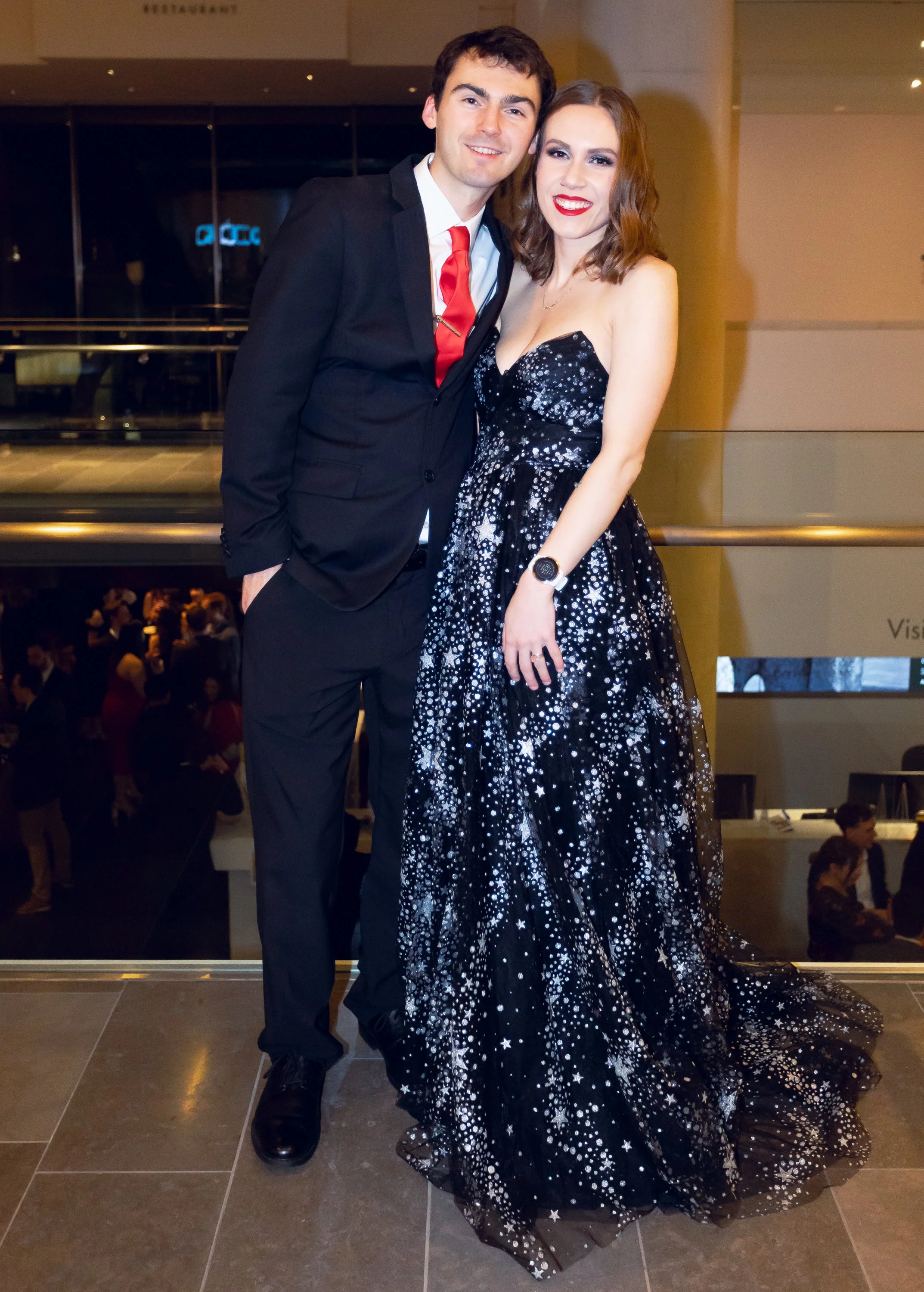 A young man in a black suit with a white shirt and red tie standing next to a young woman in a strapless black gown with white star patterns, both smiling at the camera in a public indoor space with people in the background.