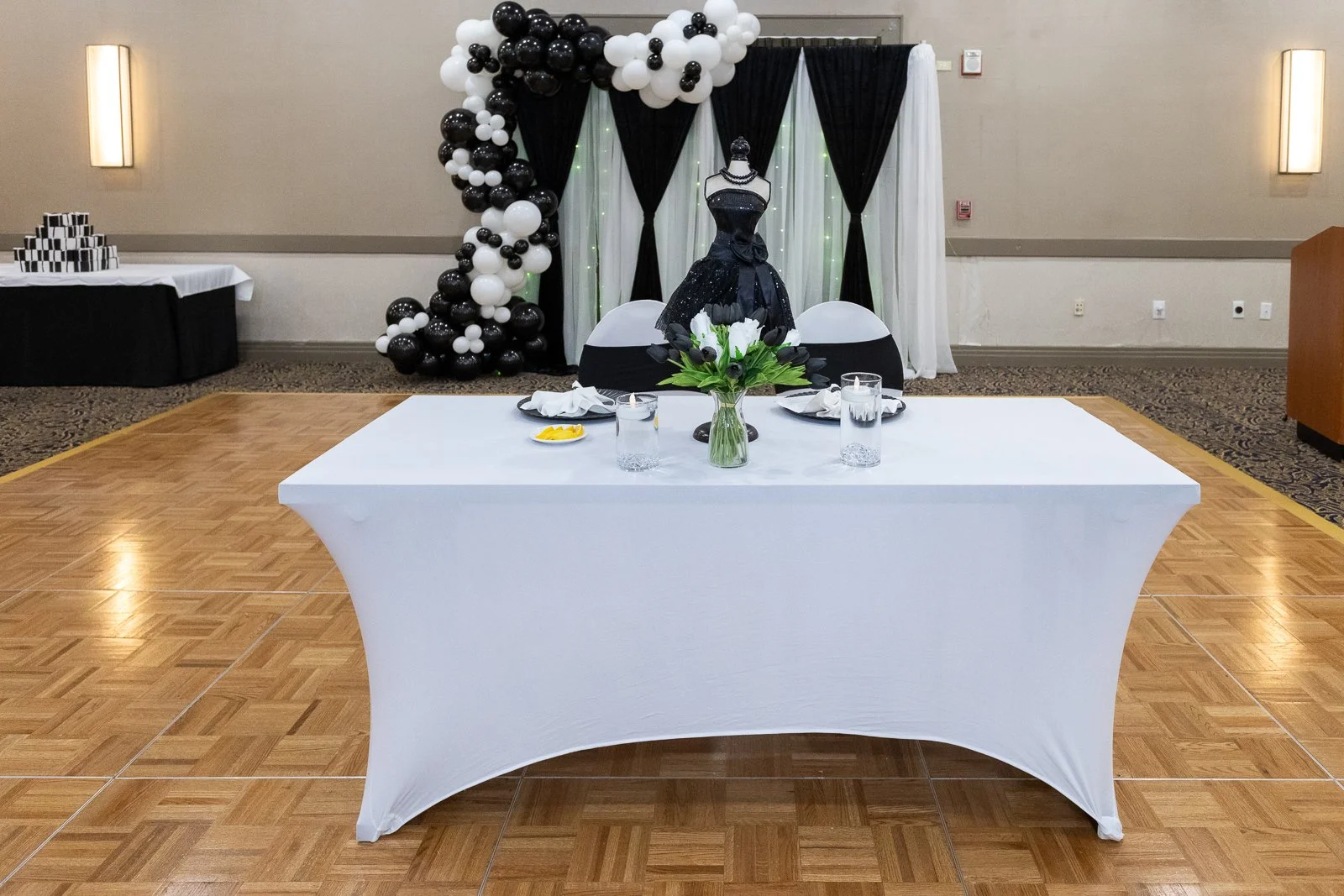 A decorated event table with a black and white theme, featuring a dress form in a black dress with a necklace, surrounded by black, white, and clear balloons, a vase of white flowers, and glasses of water on the table, set against black and white cur