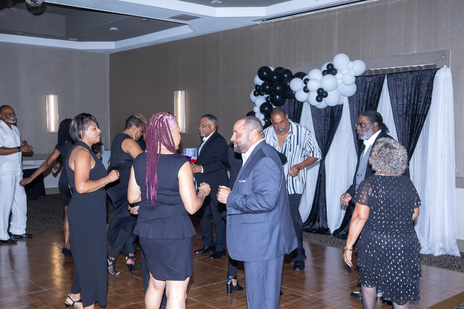 People dancing and socializing at a celebration or party in a decorated venue with black and white balloons.