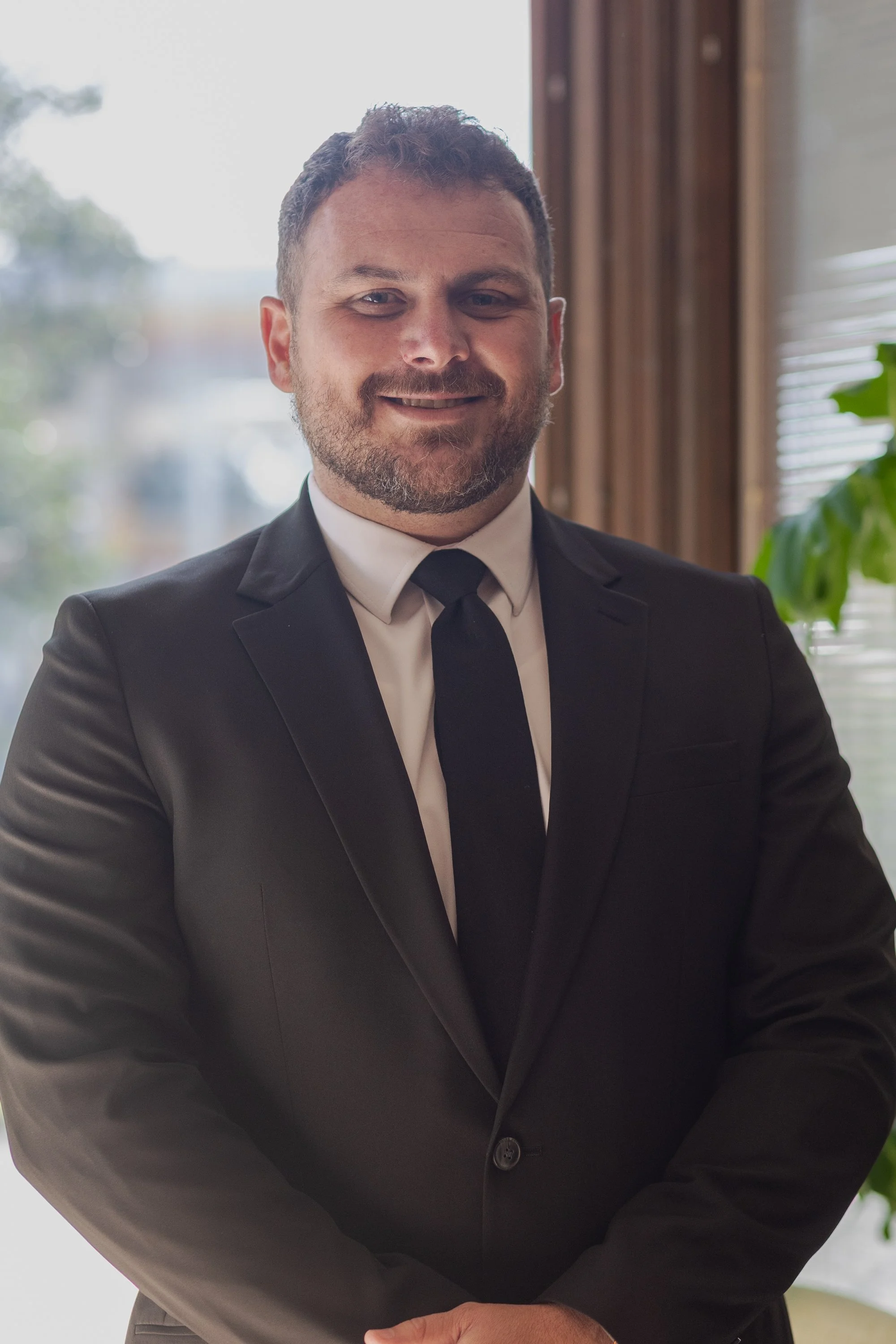 A man with short, curly brown hair and a beard, wearing a black suit with a white shirt and black tie, standing indoors near a window with partially open blinds and some greenery visible, smiling at the camera.