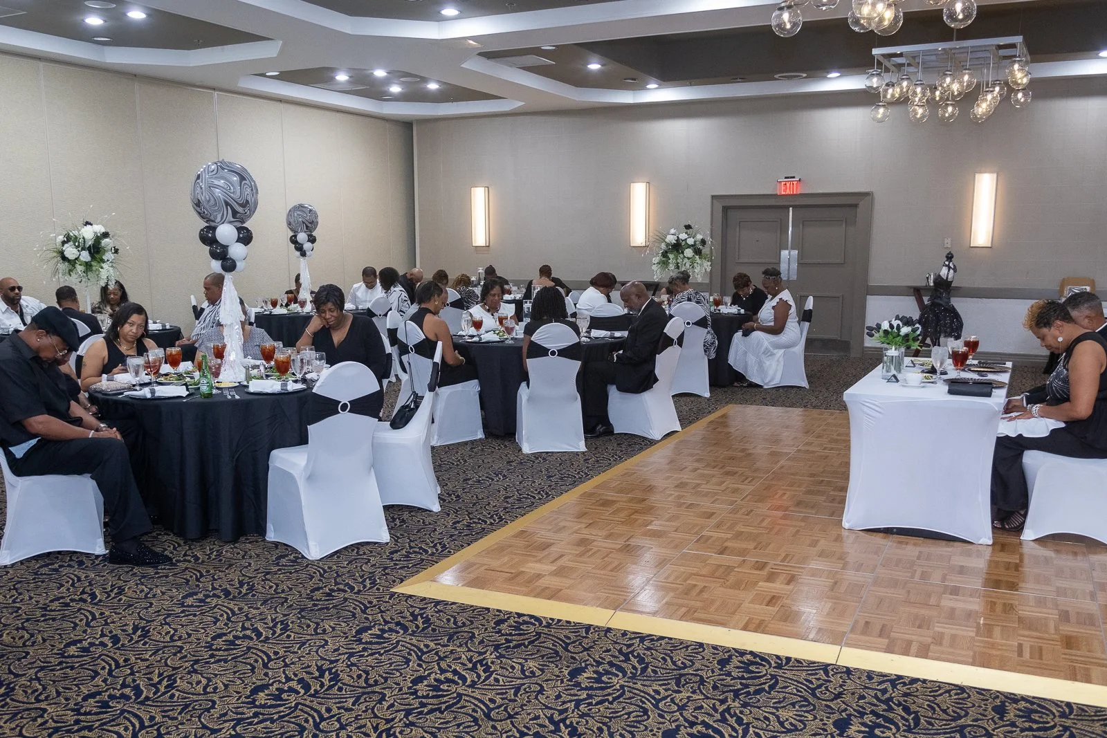 People seated at round tables with black tablecloths, white chair covers, floral centerpieces, and formal place settings in a banquet hall with a patterned carpet and a dance floor