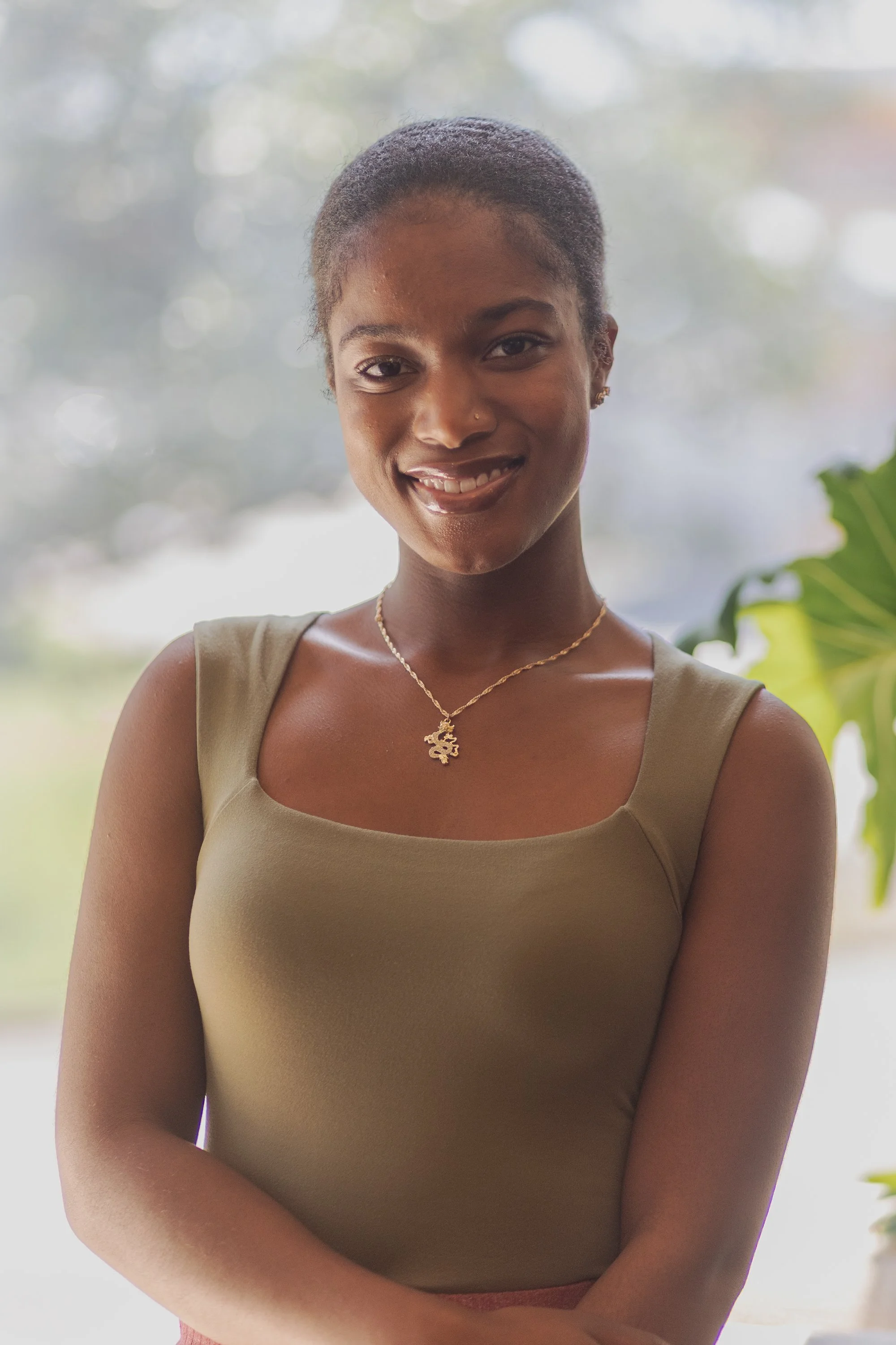 A smiling woman with short dark hair, wearing a beige sleeveless top, gold jewelry, and standing outdoors near green foliage with a blurred background.