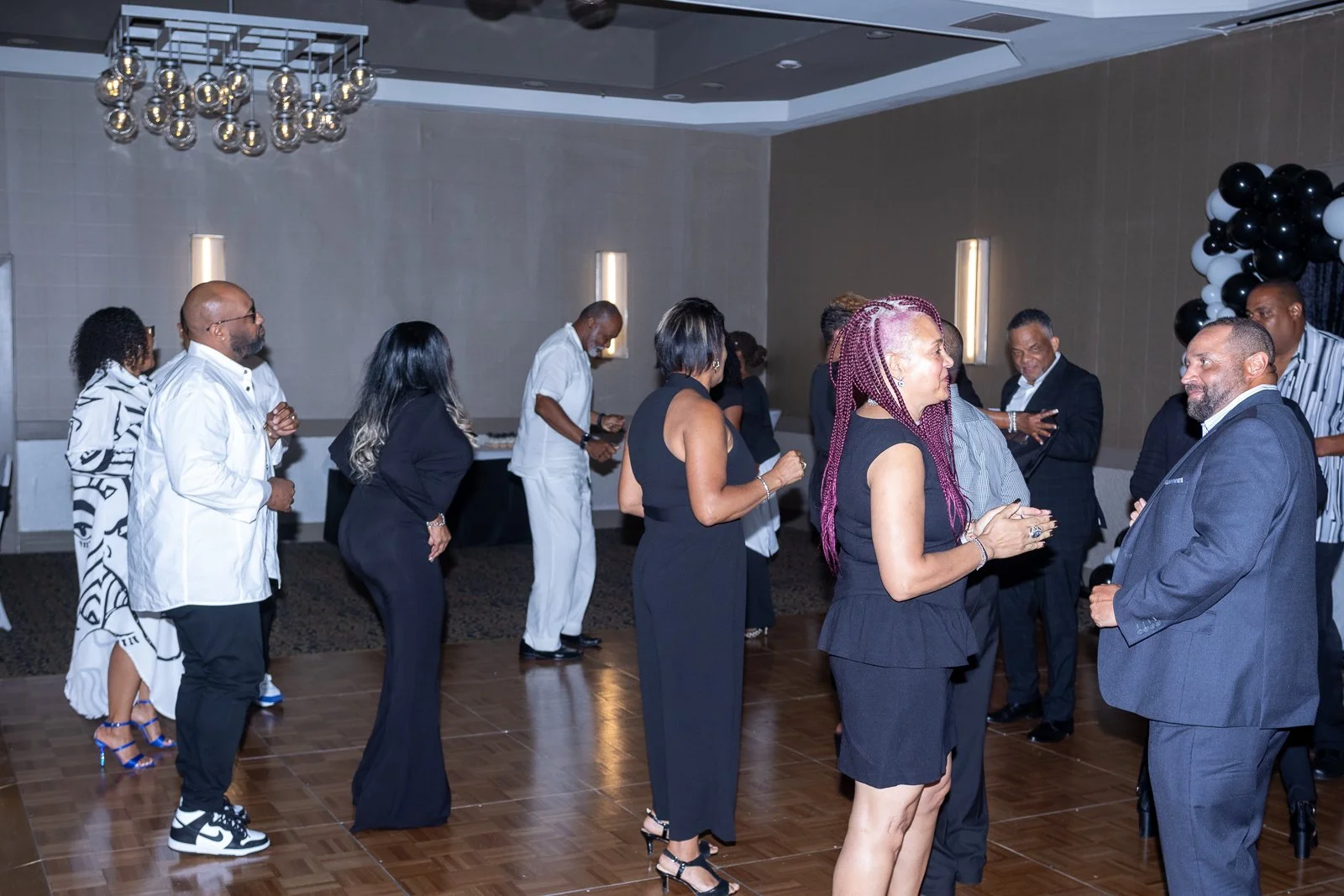 People dancing and socializing in a banquet hall with a wood dance floor, modern lighting, and black and white balloon decorations.