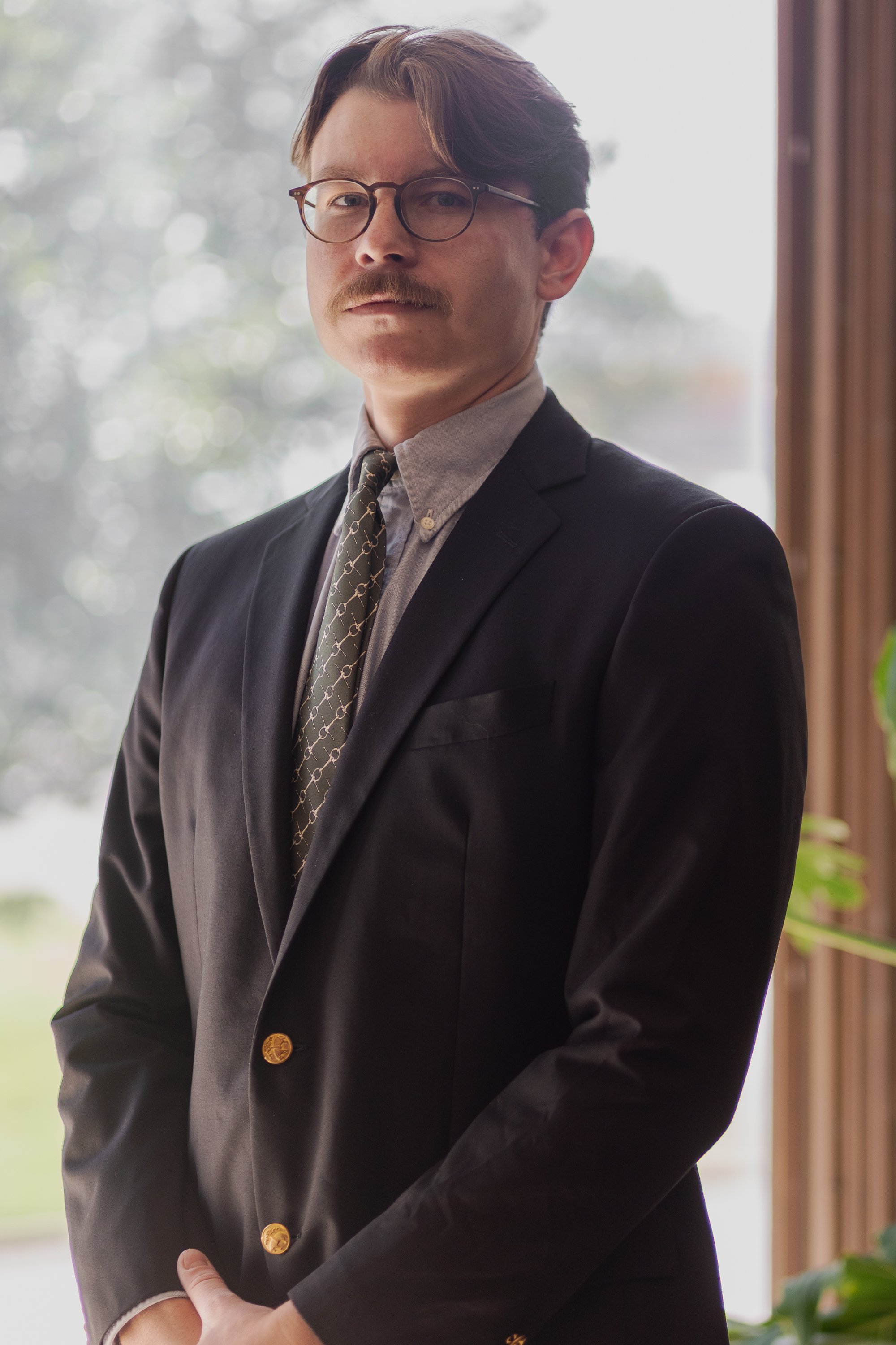 A man dressed in a suit, wearing glasses and a patterned tie, standing indoors near a window, with a blurred outdoor background.
