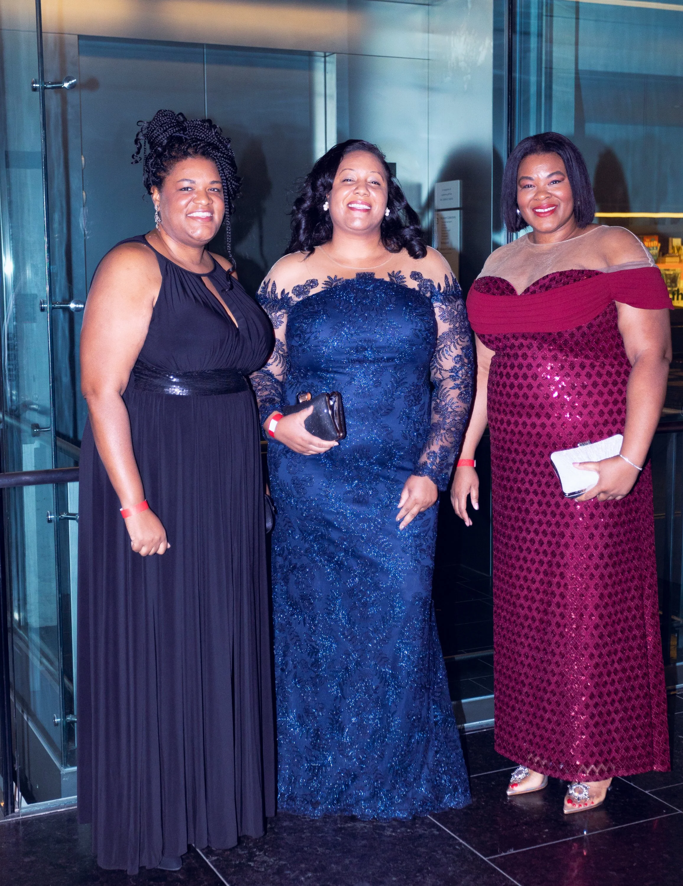 Three women in formal dresses posing together inside a building, smiling at the camera.