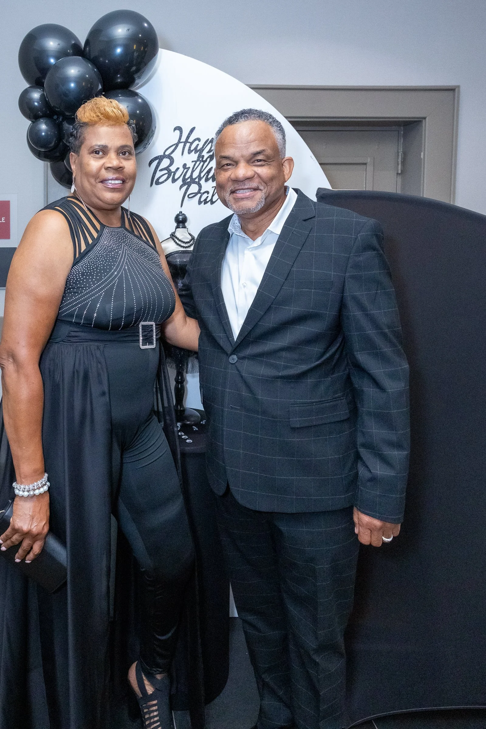 Two people dressed formally, a woman in a black dress and a man in a plaid suit, standing together at a celebration with black balloons and a white sign that says "Happy Birthday" in the background.