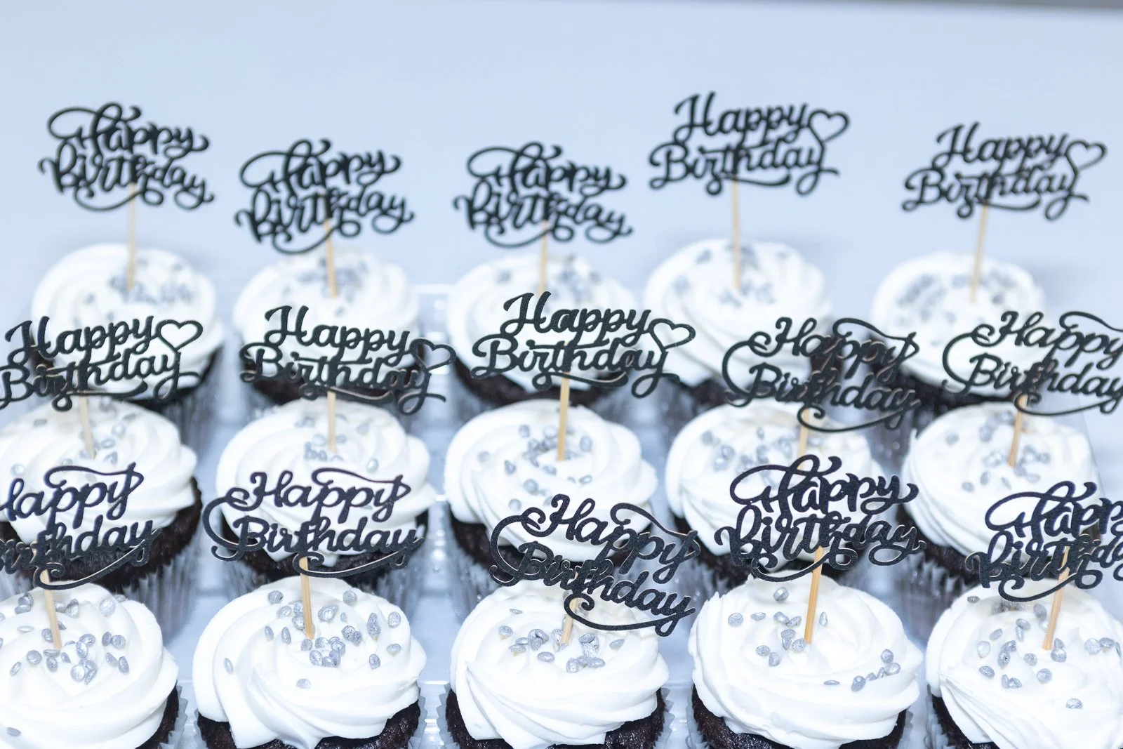 A tray of cupcakes decorated with white frosting and silver sprinkles, each with a black "Happy Birthday" topper.