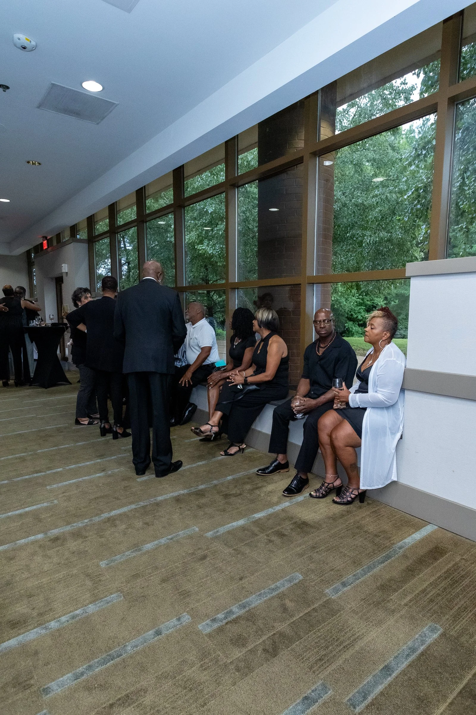 People at a social gathering or event, sitting on a bench and standing, engaging in conversations inside a room with large windows showing green trees outside.