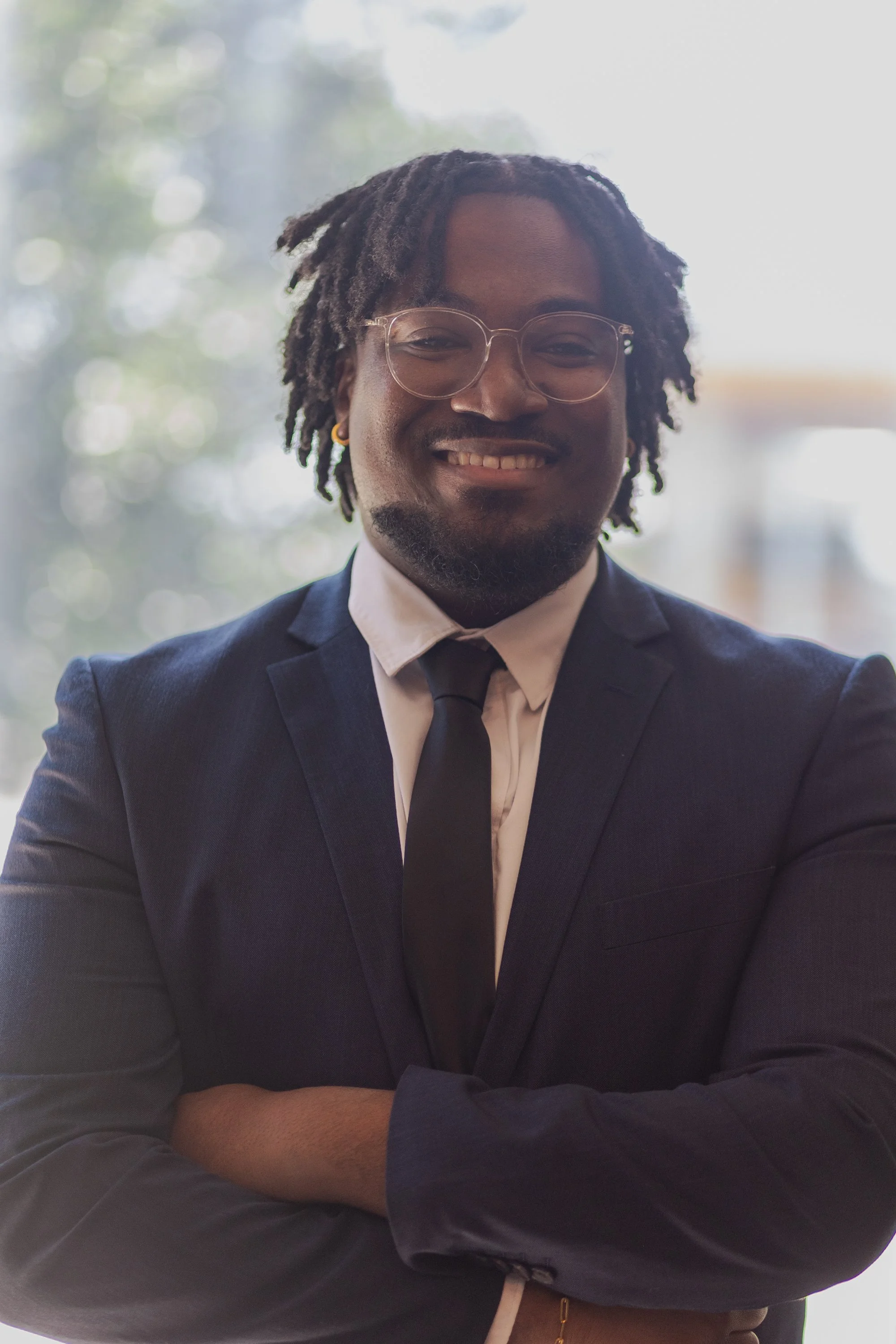 A smiling man with dreadlocks wearing glasses, a dark suit, and a white shirt with a dark tie, standing with arms crossed.