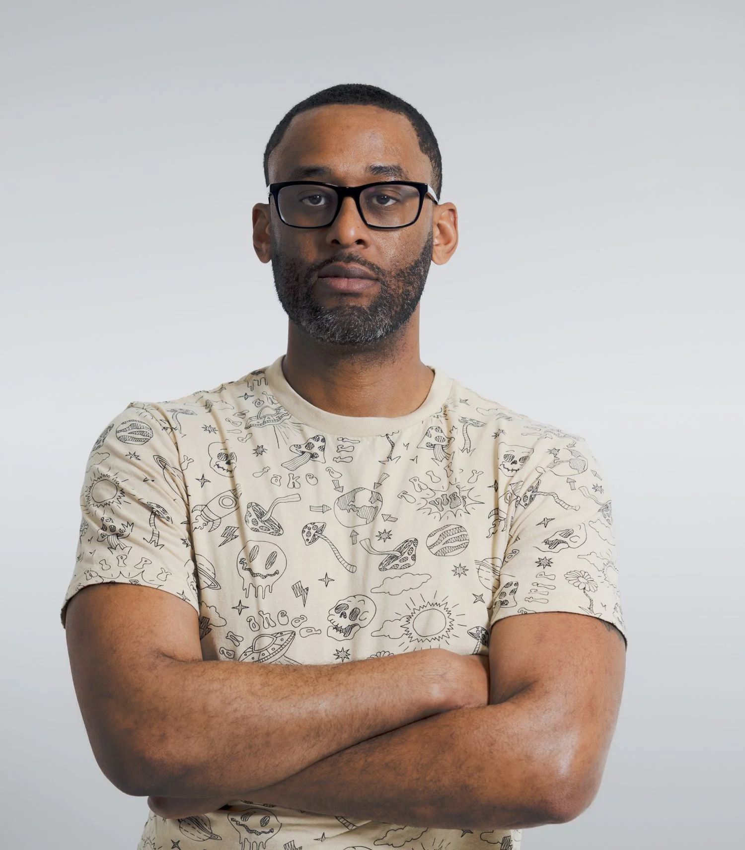 A man with glasses, a beard, and short hair crossing his arms, wearing a beige T-shirt with black line drawings of skulls, planets, and other doodles, standing against a plain light gray background.