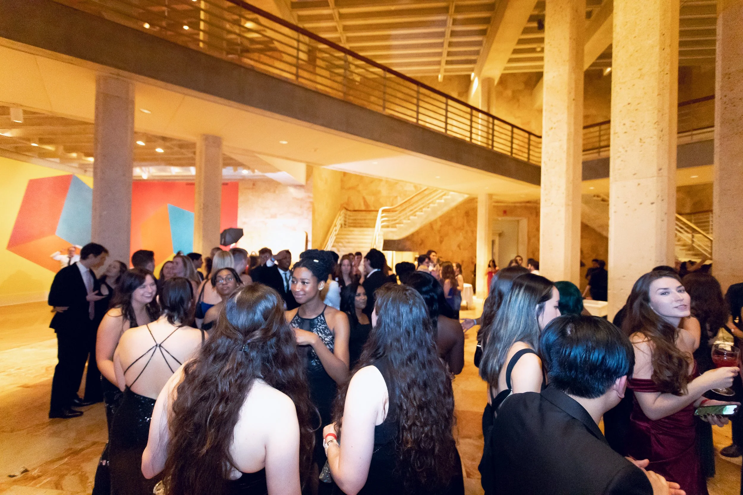 A large crowd of people dressed in formal attire gathered inside a spacious, well-lit hall with high ceilings, a staircase, and colorful abstract art on the wall.