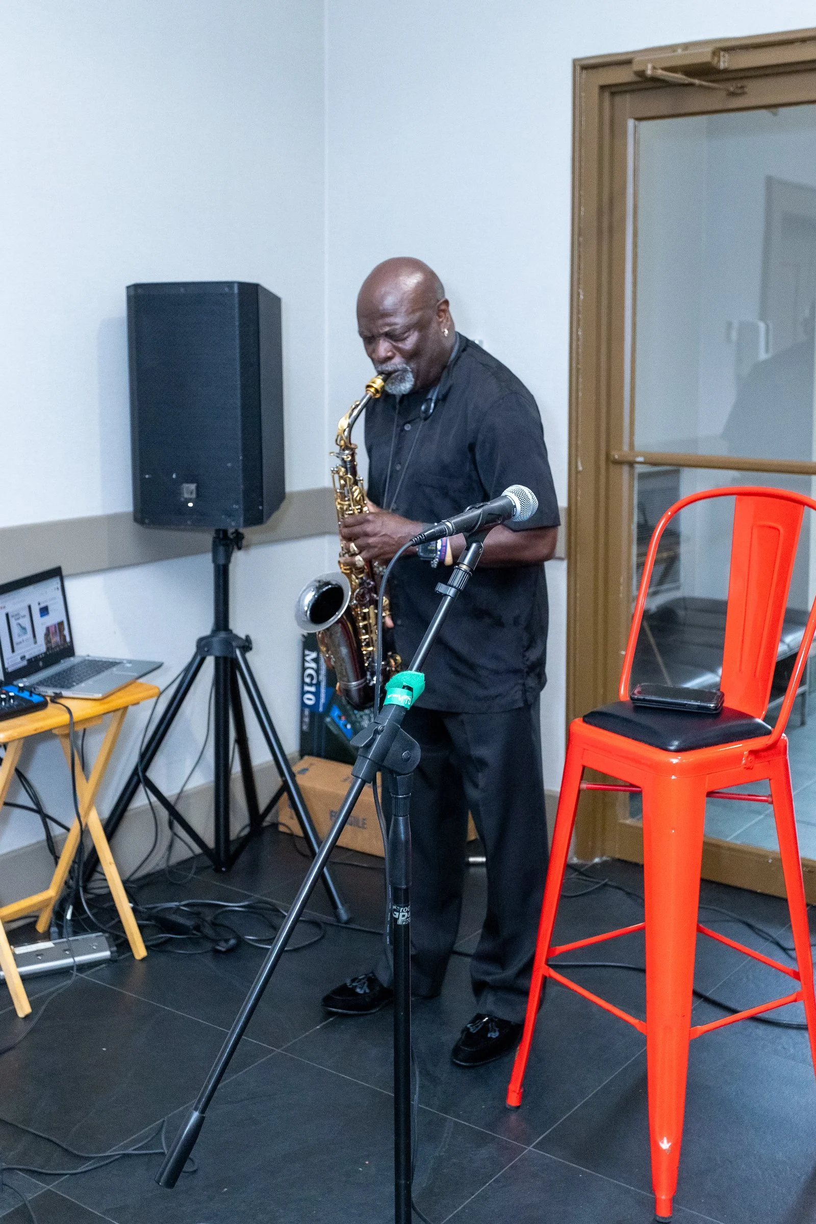A man playing a saxophone in a room with sound equipment and a bright orange chair.