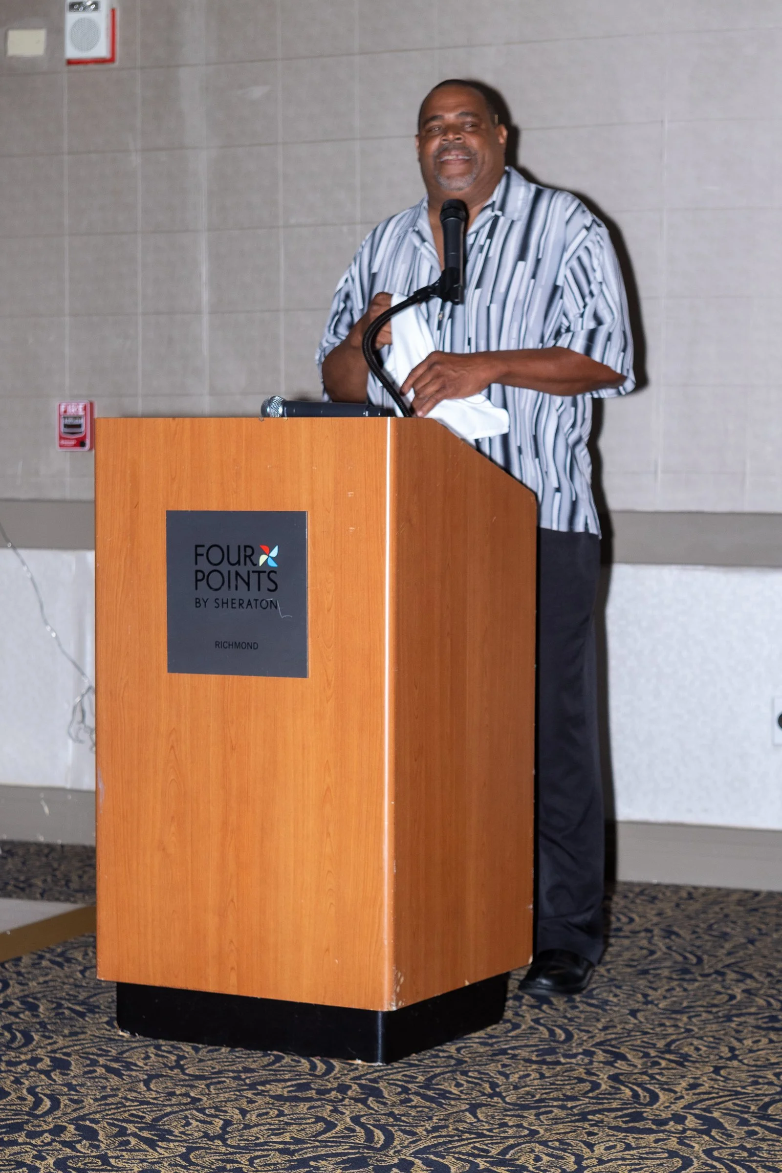 A man standing behind a podium at a conference or event, wearing a striped shirt, with a microphone in hand, in a hotel conference room.