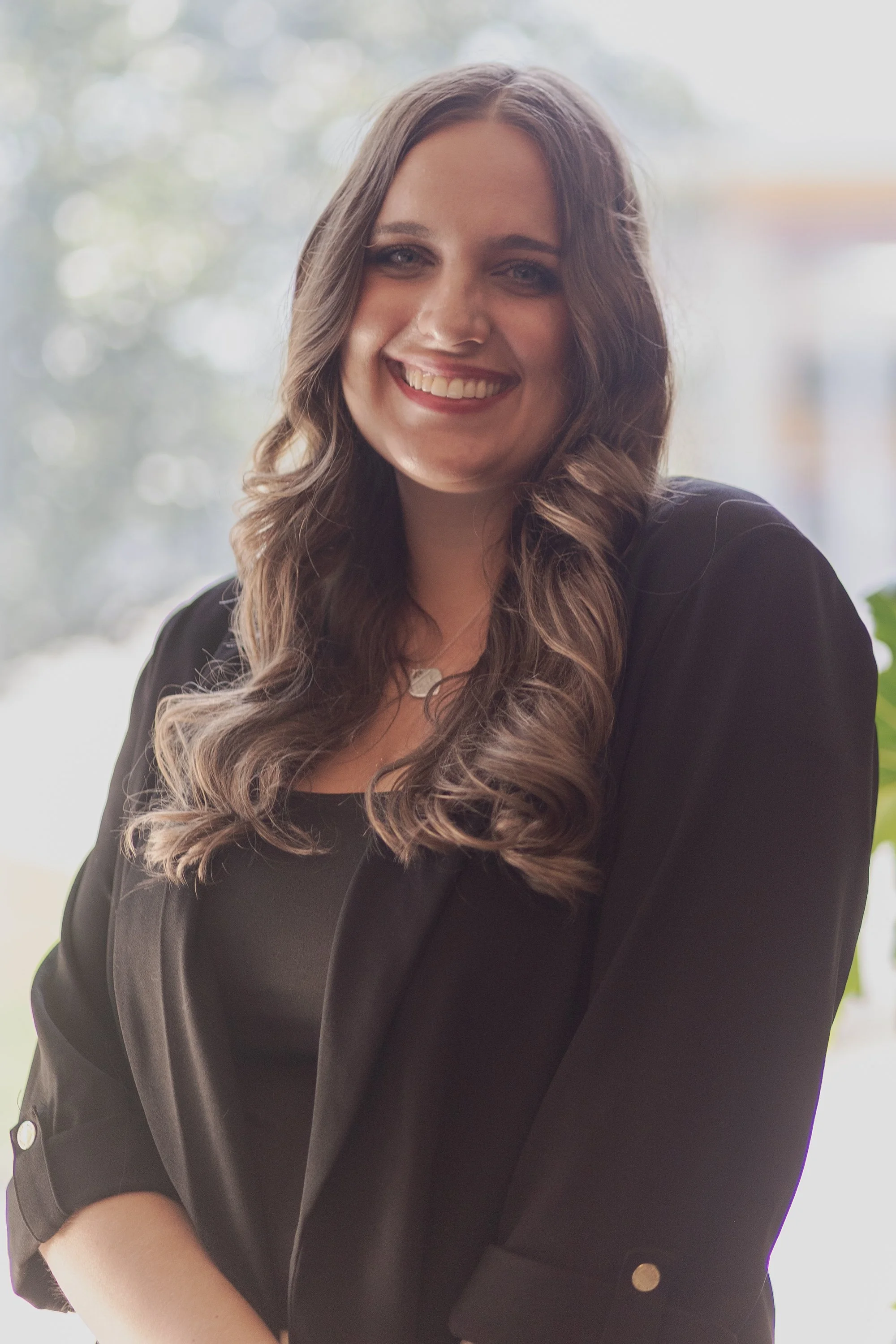 A woman with long, wavy brown hair wearing a black blazer and smiling, standing with a bright, blurred background.