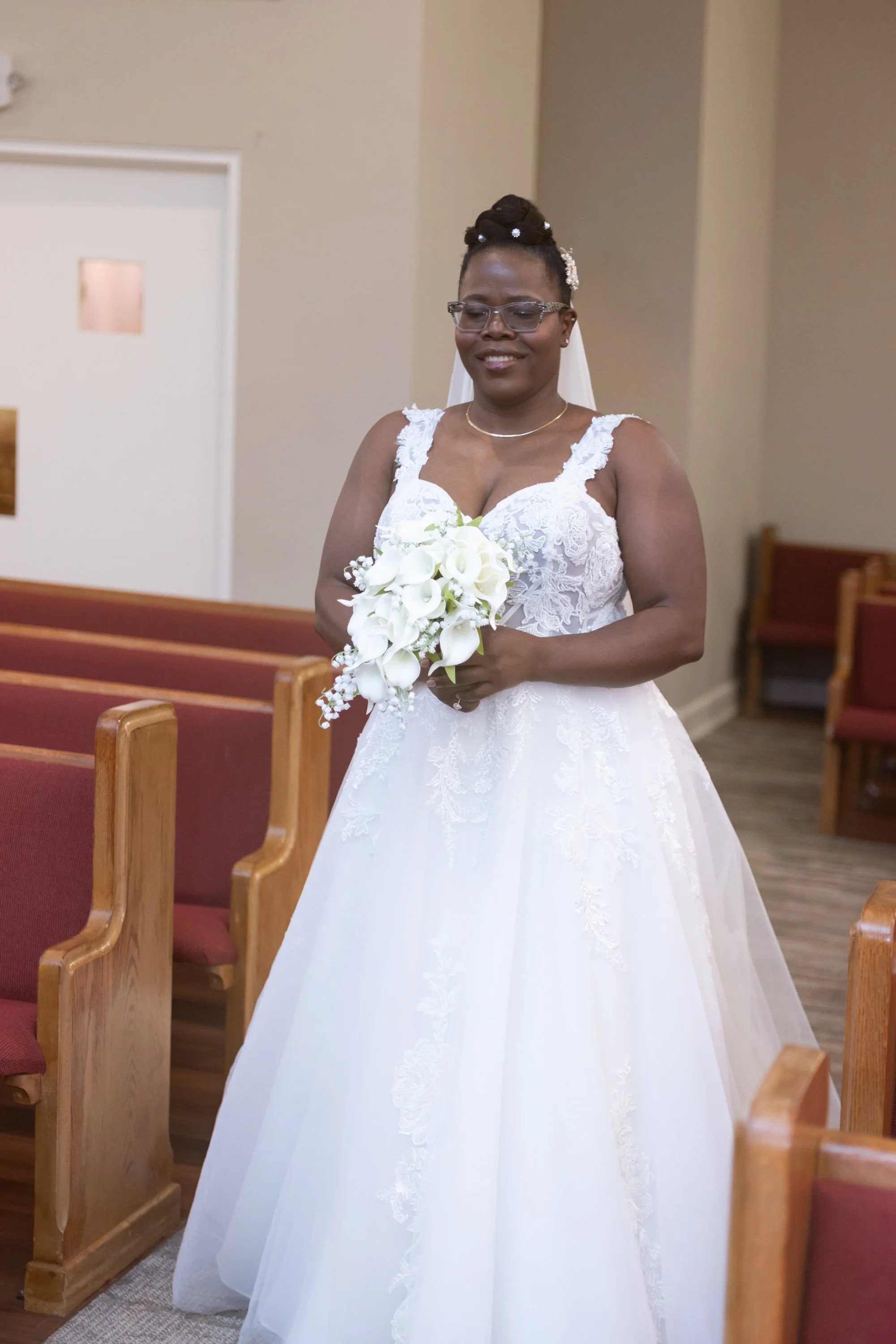 A bride in a white lace wedding dress holding a bouquet of calla lilies, standing inside a church.