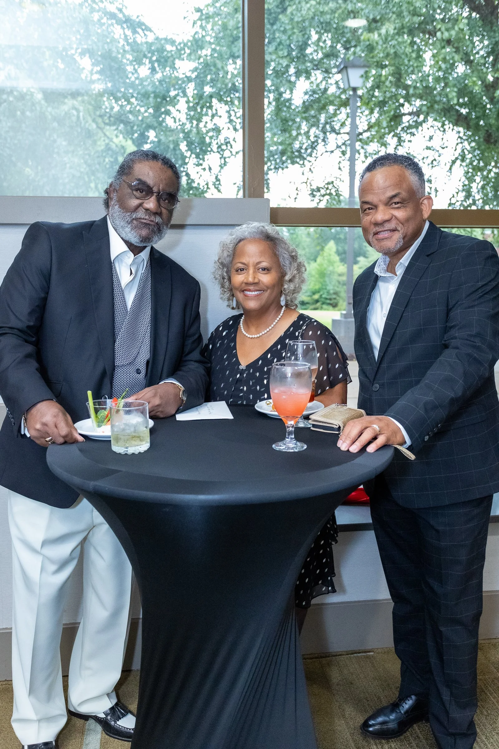 Three well-dressed people, two men and one woman, standing around a black round table with drinks, in a room with large windows showing trees outside.