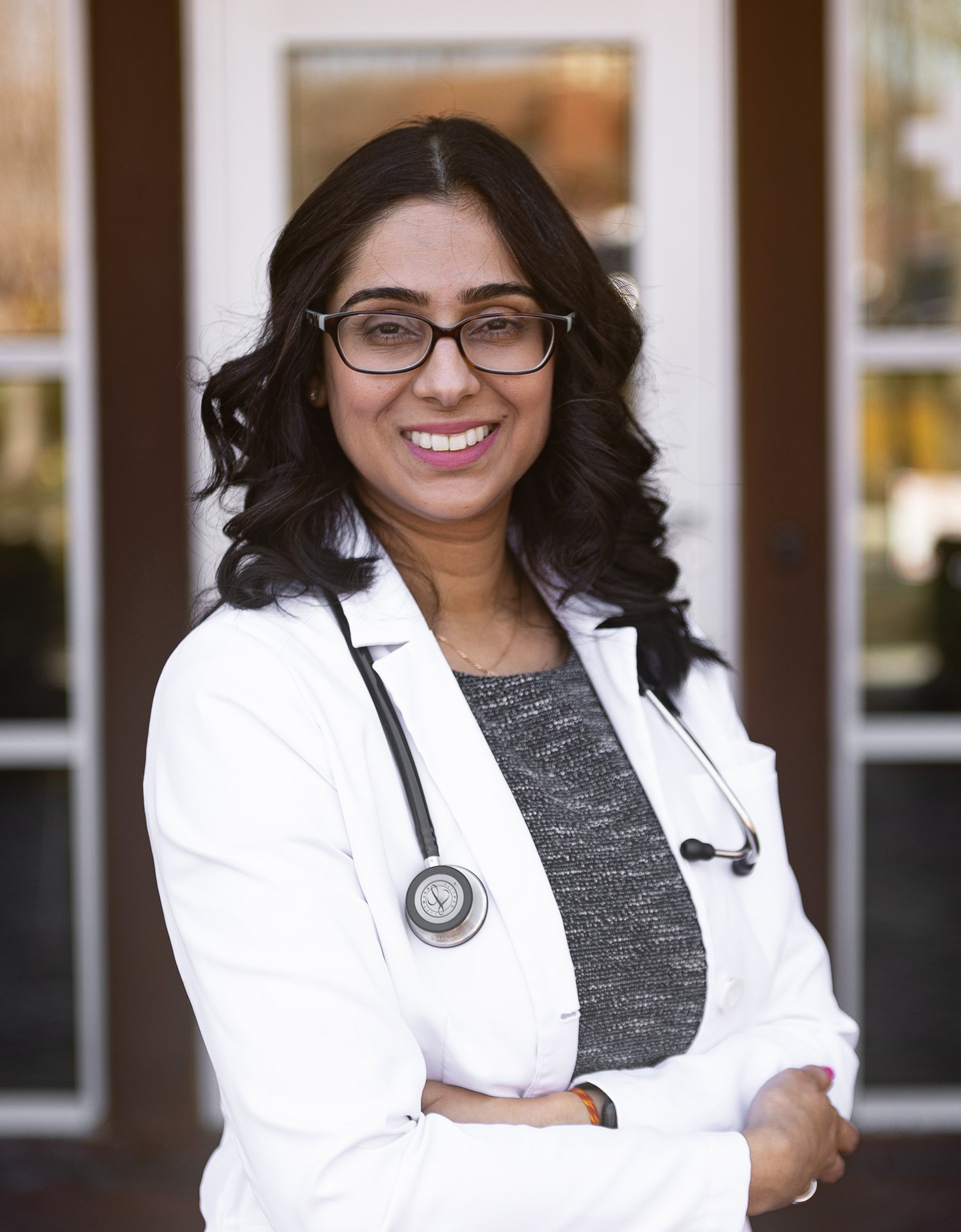 A female doctor with dark hair, glasses, and a stethoscope around her neck standing outdoors, smiling with her arms crossed.