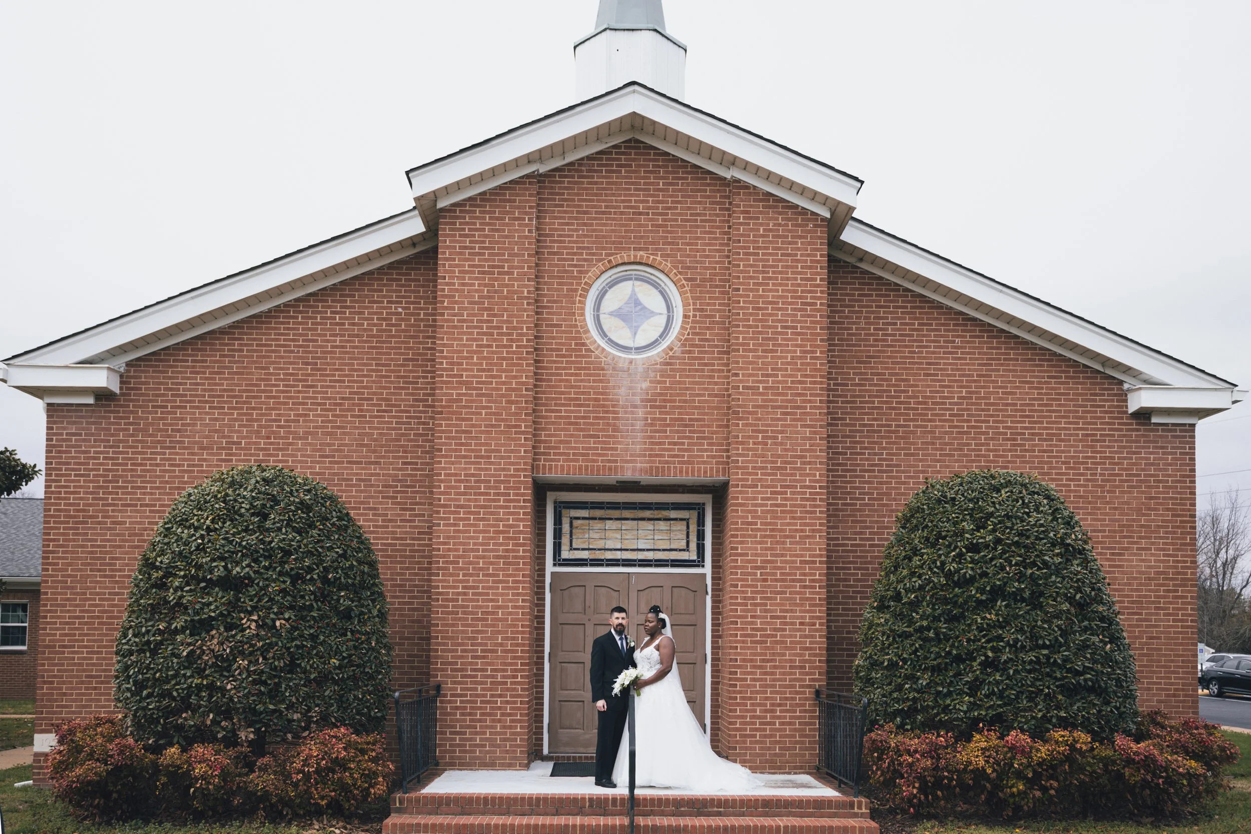 A newlywed couple dressed in wedding attire standing in front of a brick church building, holding a bouquet of flowers, with two large round bushes and flowering shrubs in the foreground.