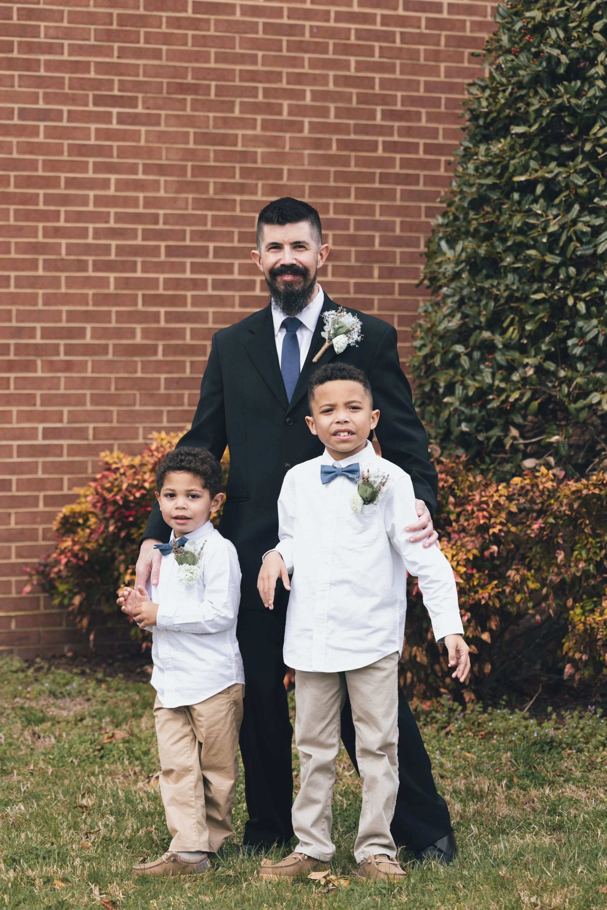 A man with a beard and mustache wearing a black suit with a white shirt, navy tie, and boutonniere standing outdoors in front of a brick wall and foliage. He is accompanied by three young boys dressed in white shirts, khaki pants, and bow ties, stand