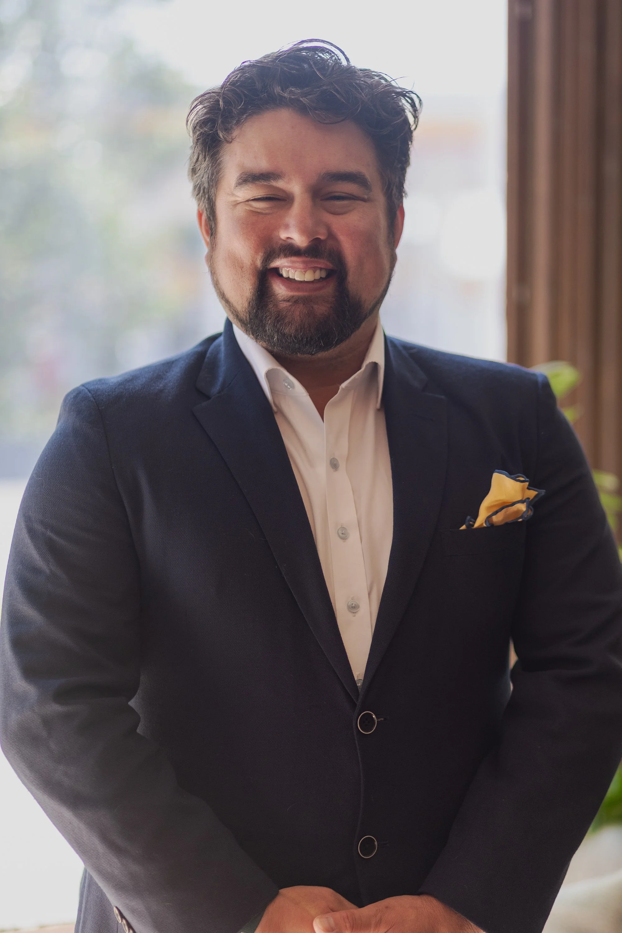 A man wearing a dark suit with a yellow pocket square, white shirt, and a blue tie, smiling with his eyes closed, indoors near a window with blurred outdoor scenery.