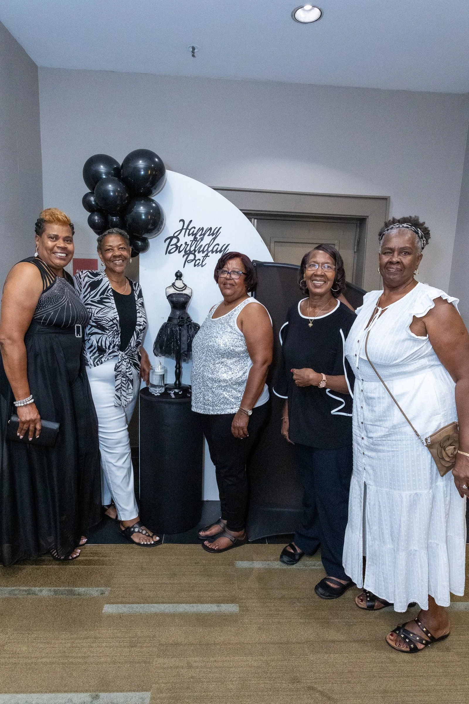 Group of six women standing together in front of a birthday-themed backdrop with black balloons and a mannequin display, celebrating a birthday with the message 'Happy Birthday Pat' written on the backdrop.