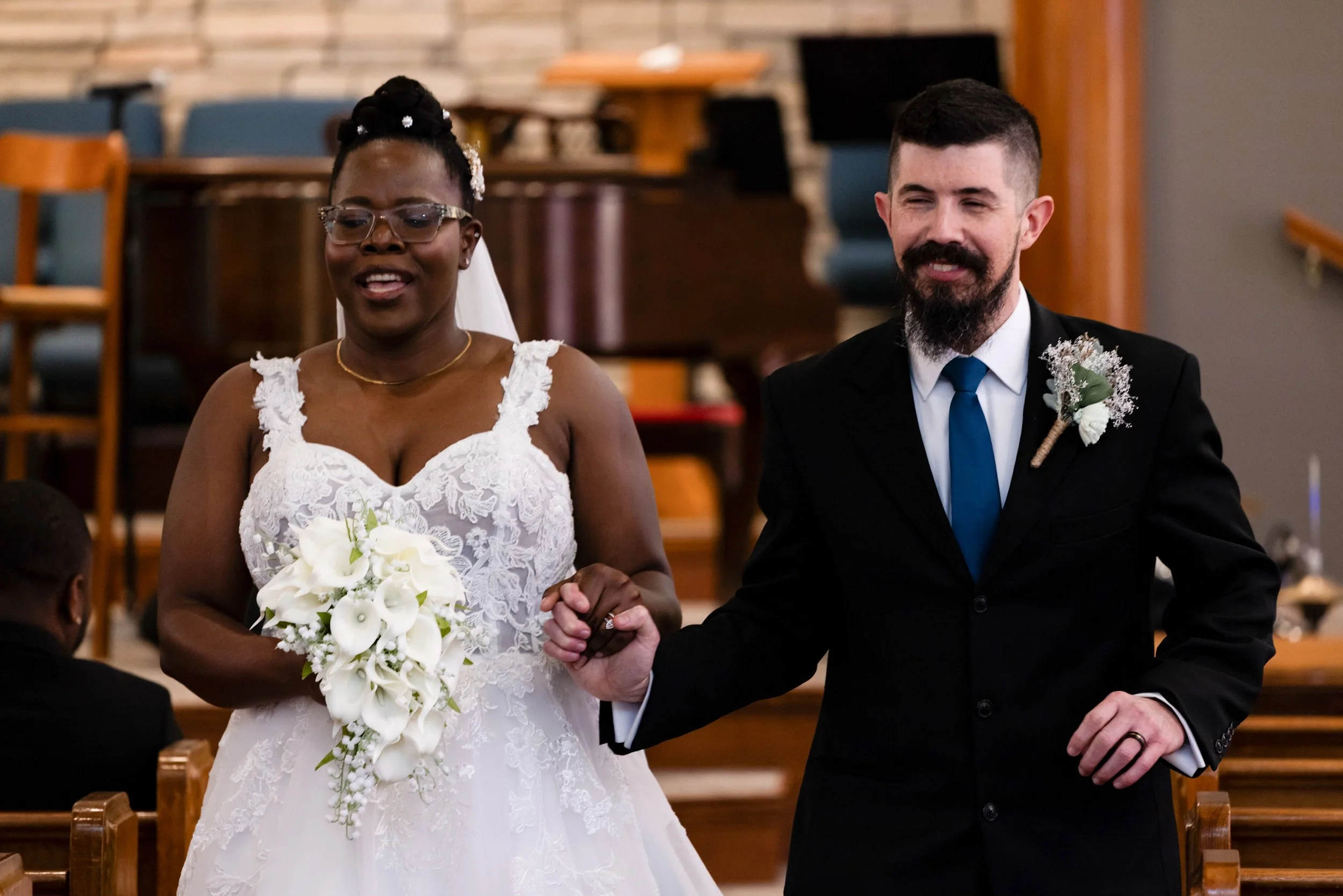 A bride in a white lace wedding dress holding a bouquet of white flowers, wearing glasses and a veil, stands next to a groom in a black suit, white shirt, and blue tie, inside a church during a wedding ceremony.
