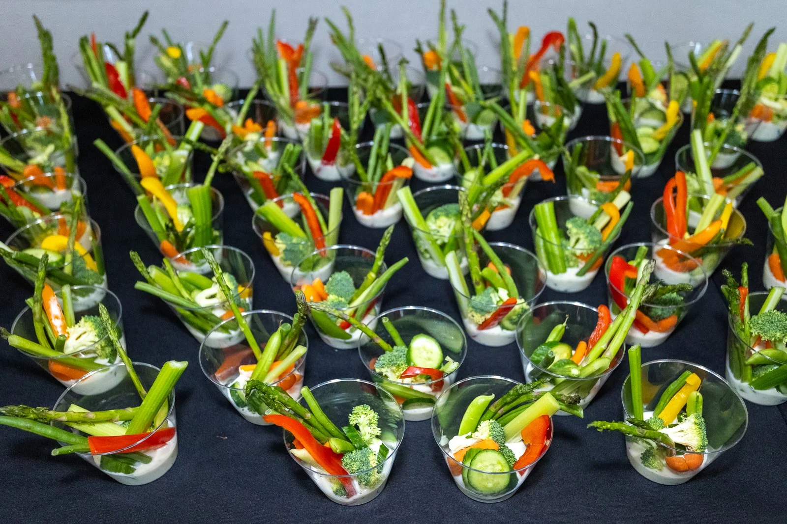 Multiple small clear cups filled with fresh vegetable sticks including broccoli, celery, red and yellow bell peppers, cucumber, and asparagus, arranged on a dark surface.
