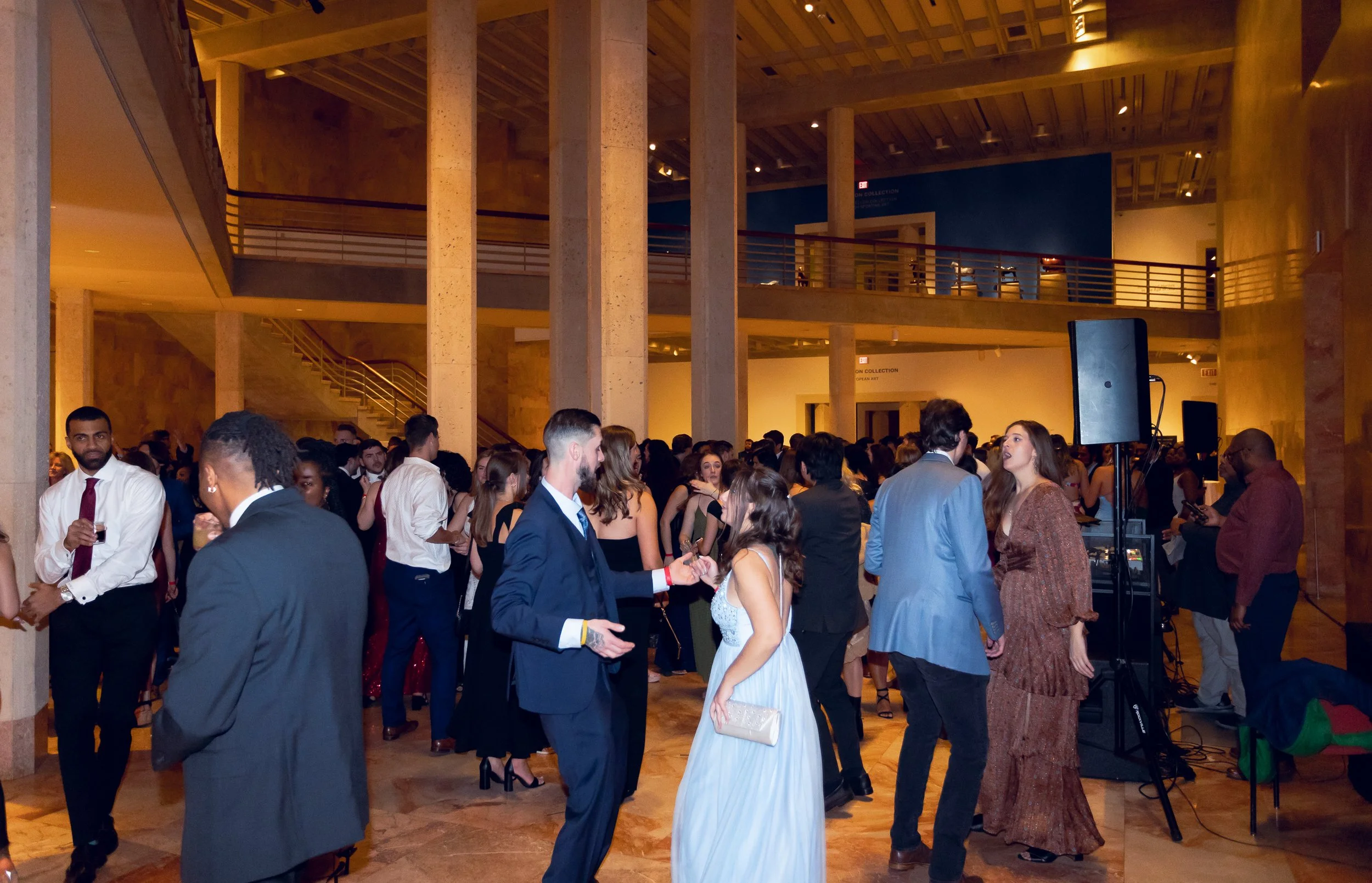 Indoor event with many people dressed in formal attire, mingling and talking in a spacious, well-lit hall with wooden walls and balconies.