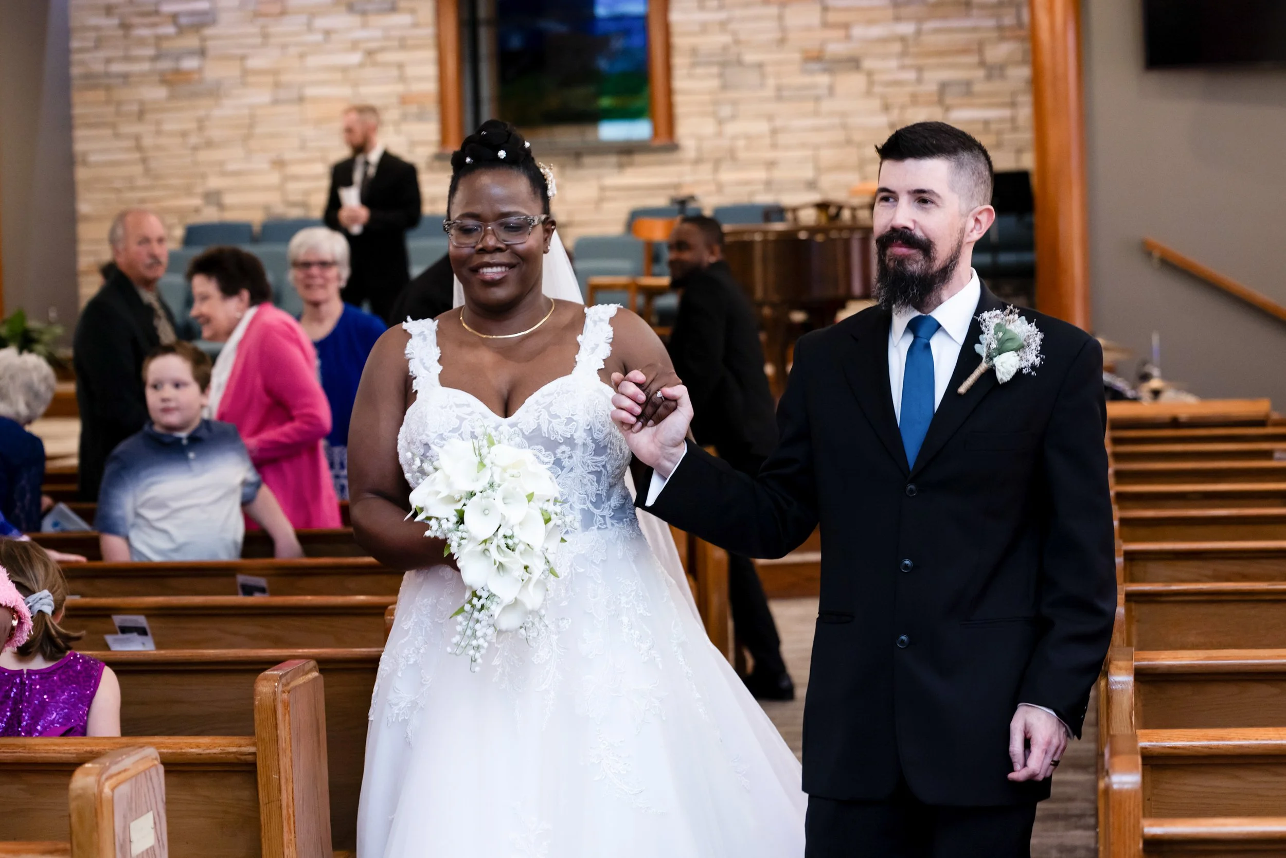 A bride and groom holding hands during their wedding ceremony inside a church, with wedding guests seated in the background.