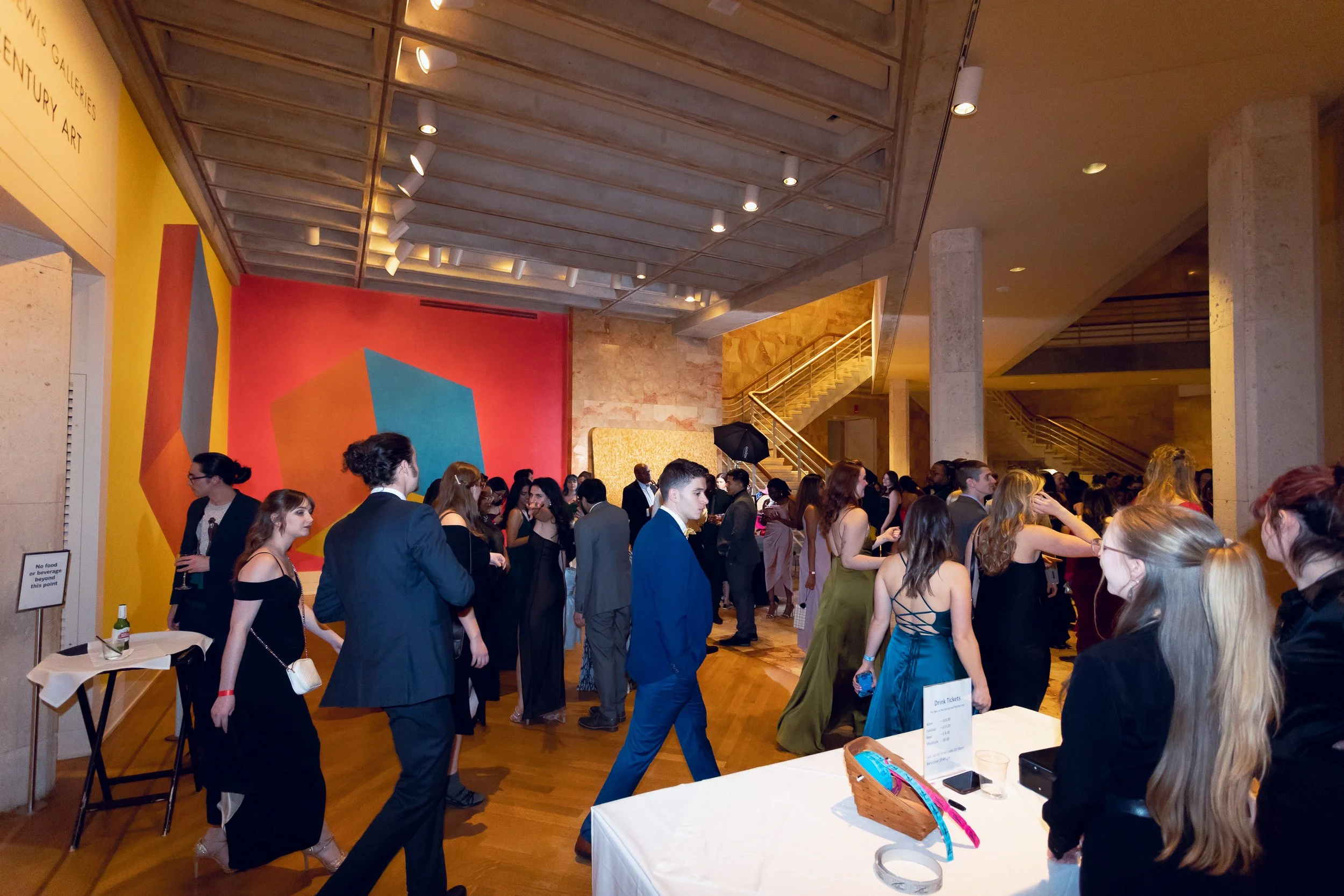A busy art gallery or museum with people dressed in formal attire attending an event. There is a colorful geometric mural on the large wall to the left, spotlights on the ceiling, a staircase in the background, and a table with drink tickets and a ba