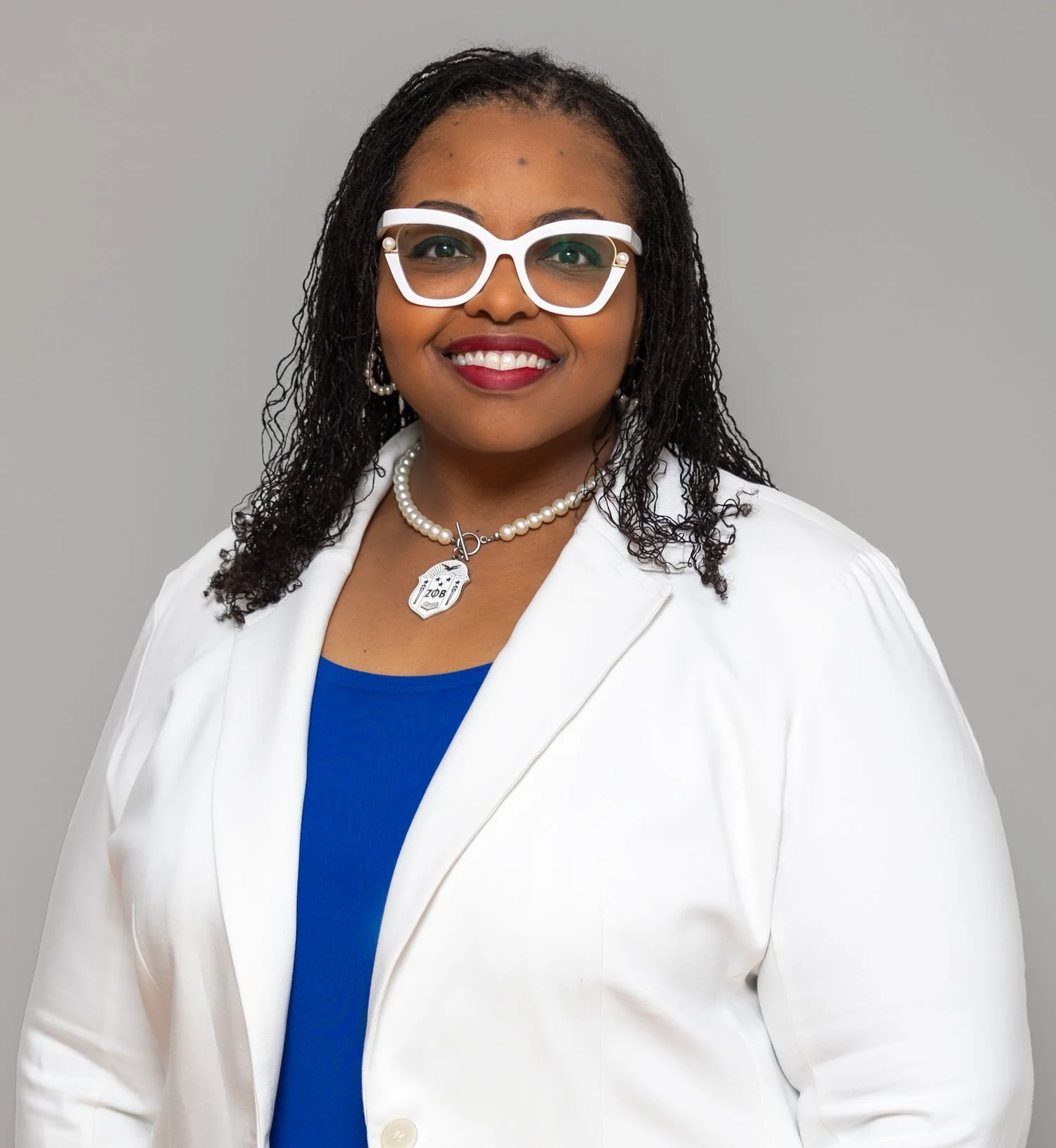 Portrait of an African American woman with curly hair wearing large white glasses, pearl earrings, a pearl necklace with aDelta Zeta badge, a white blazer, blue top, and red lipstick, smiling against gray background.