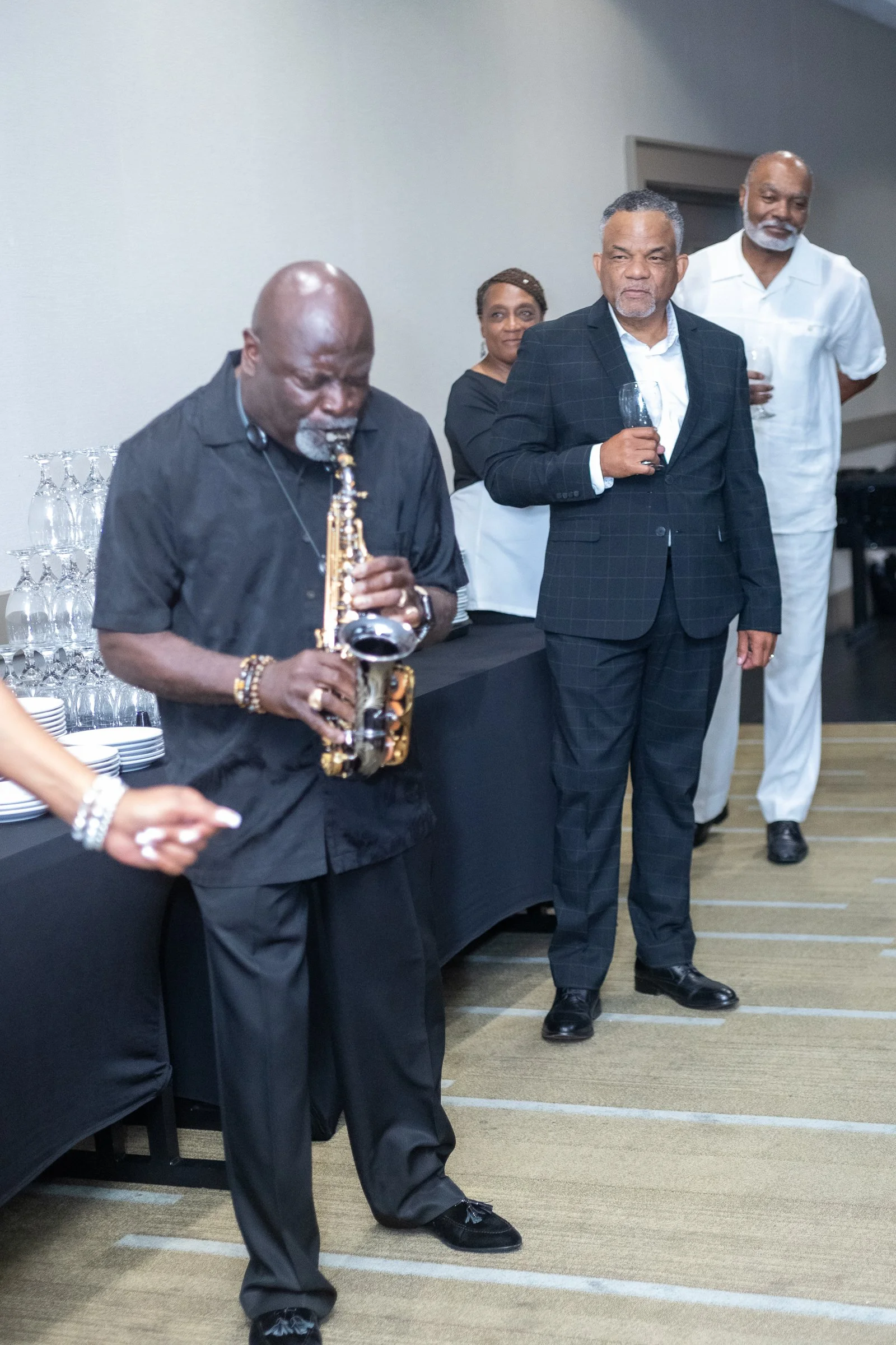 Group of African American adults dressed in formal attire at a social event, with some holding drinks, standing on a patterned carpet.