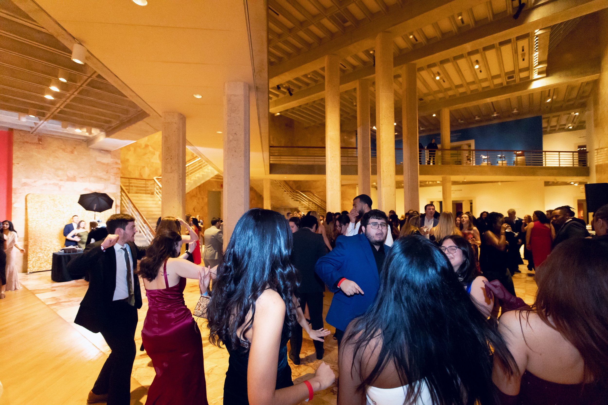 A large group of people dressed in formal attire, socializing and dancing at an indoor event with high ceilings, wooden beams, and warm lighting.