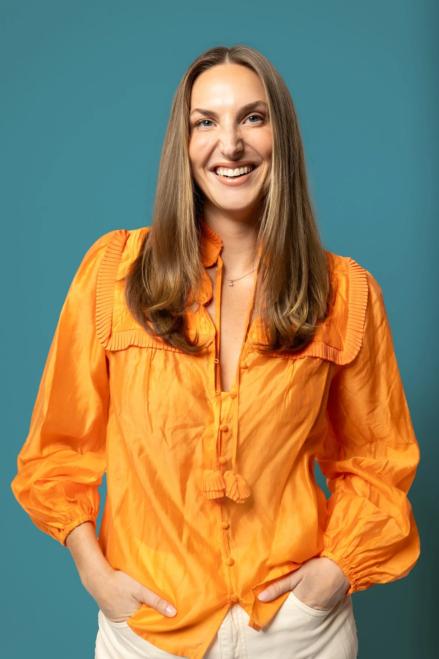 A woman with long brown hair smiling, wearing an orange silky blouse with pleated details and beige pants, standing against a teal background.