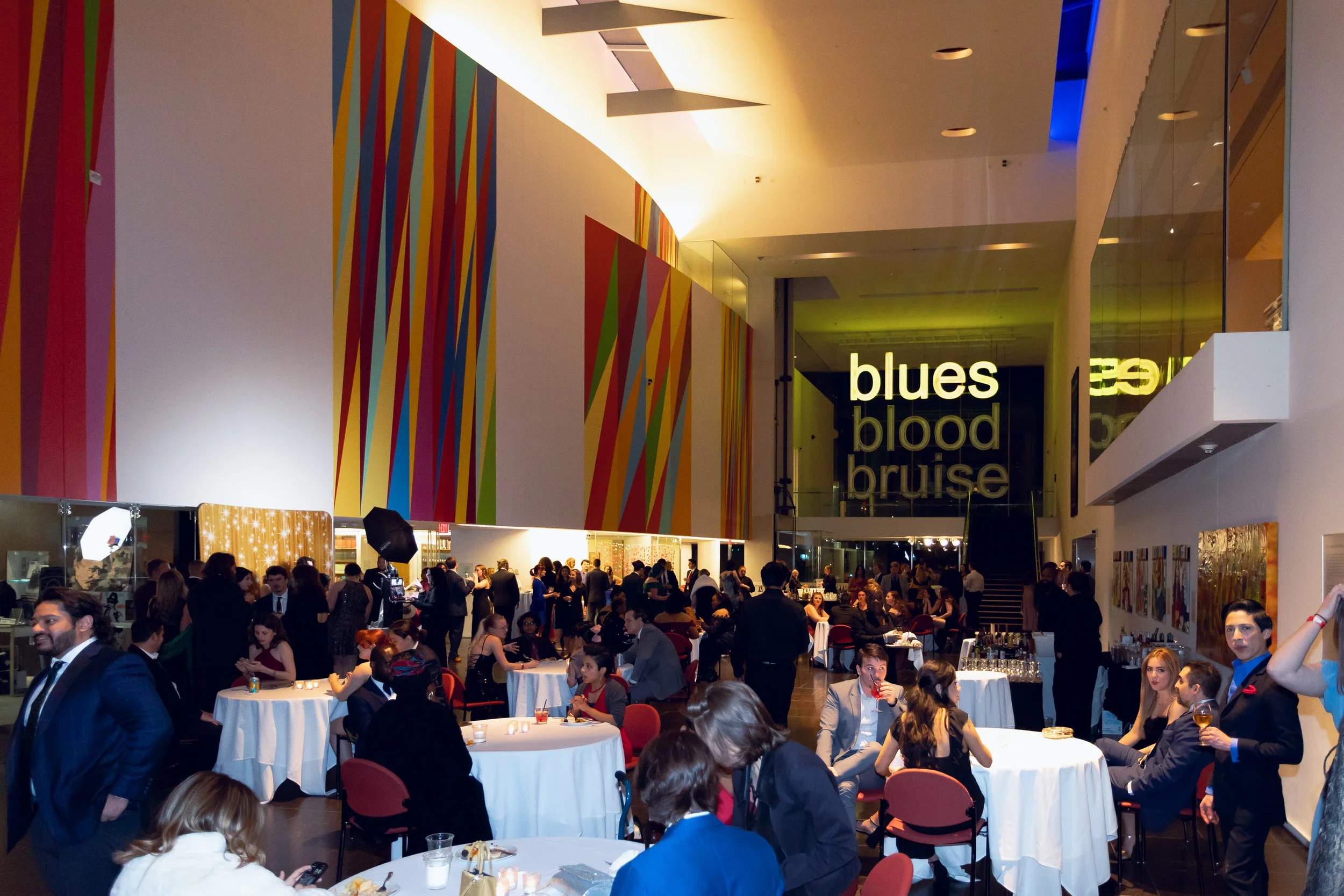 A large indoor event with many people seated at round tables covered with white tablecloths, some standing and others seated and dining. The background features a colorful vertical striped wall and signage with the words 'blues blood bruise'. The spa