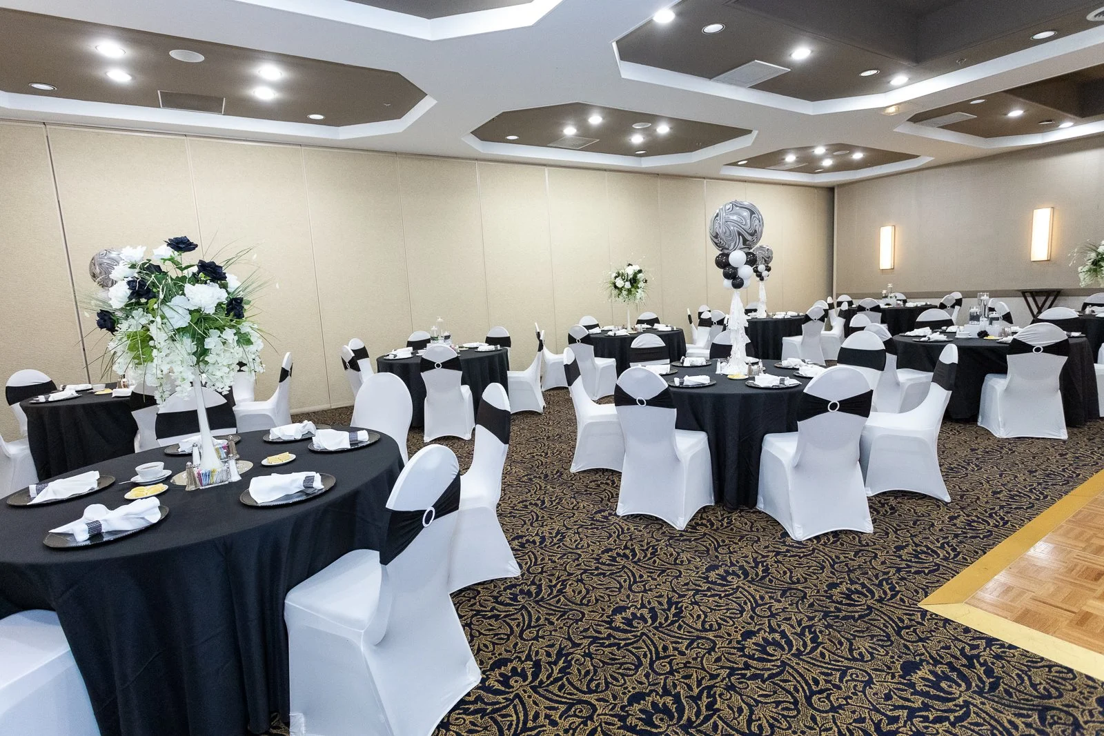 Banquet hall decorated for an event with round tables covered in black tablecloths, white chairs with black sashes, floral centerpieces, and balloon decorations in black, white, and marble patterns.