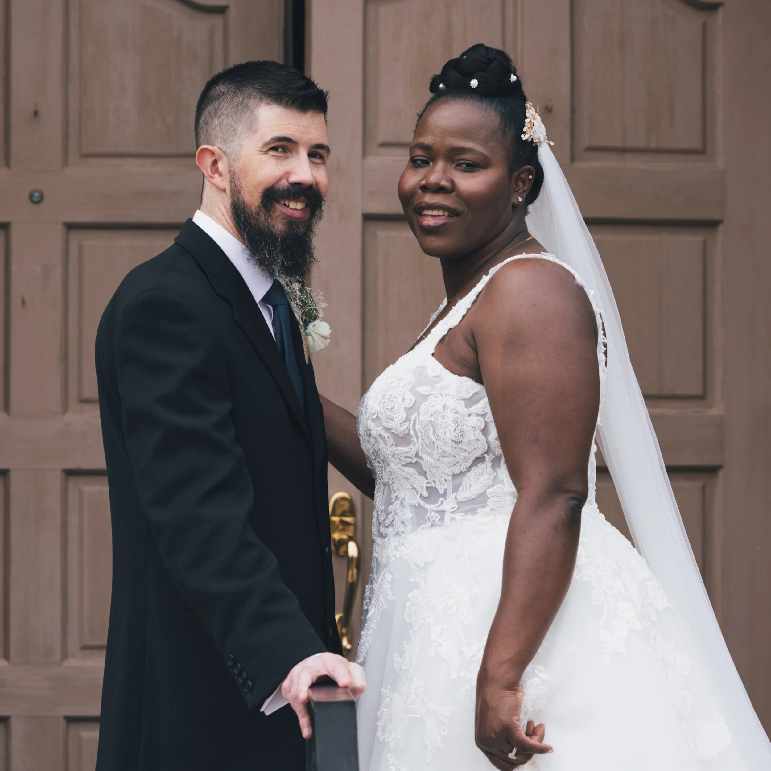 A couple dressed in wedding attire holding hands, standing outside in front of a wooden door.