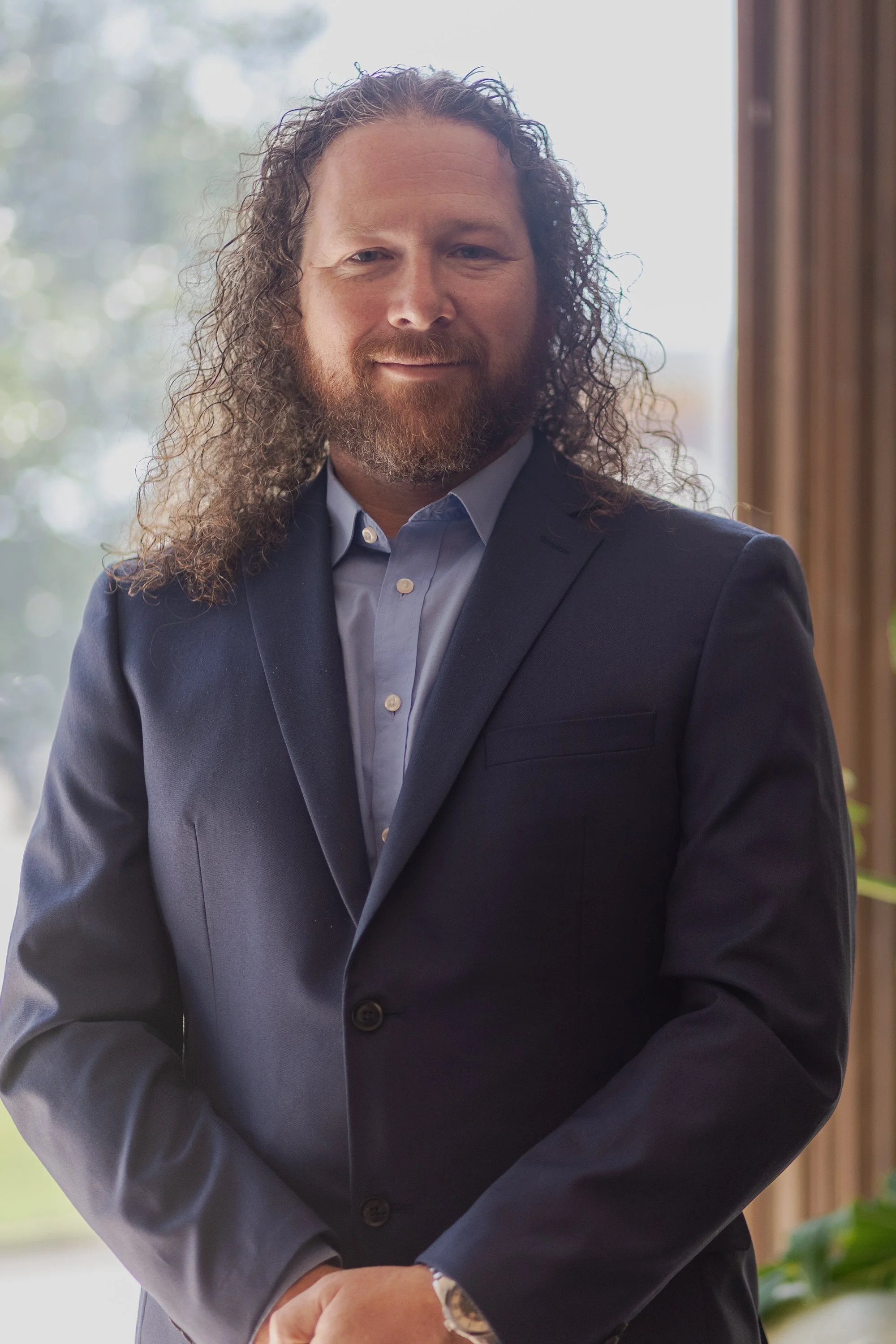 A man with long curly hair and a beard wearing a dark suit and light blue shirt standing indoors in front of large windows with natural light.