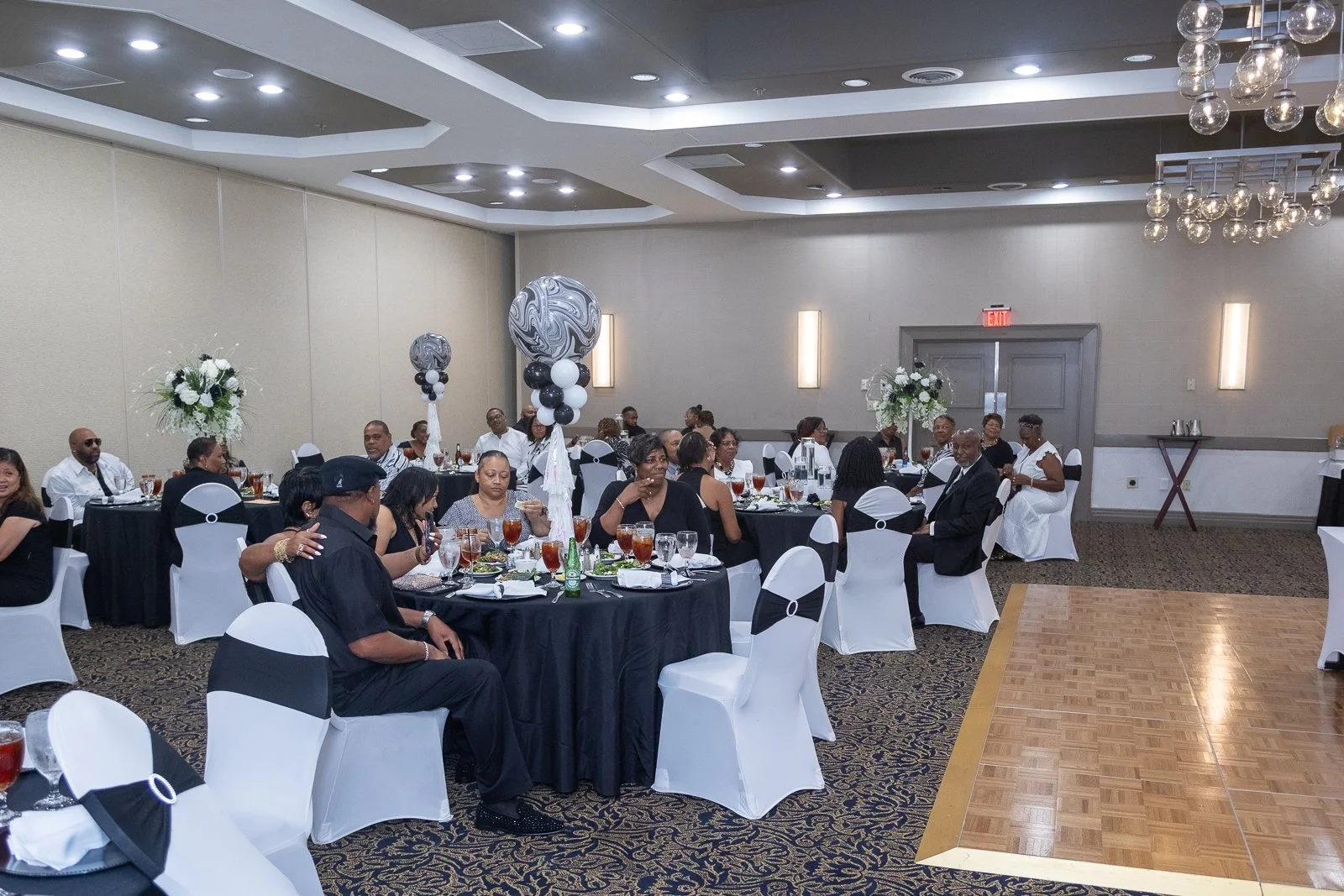 People seated at round tables in a banquet hall for a formal event with black and white decorations, floral centerpieces, and balloons.