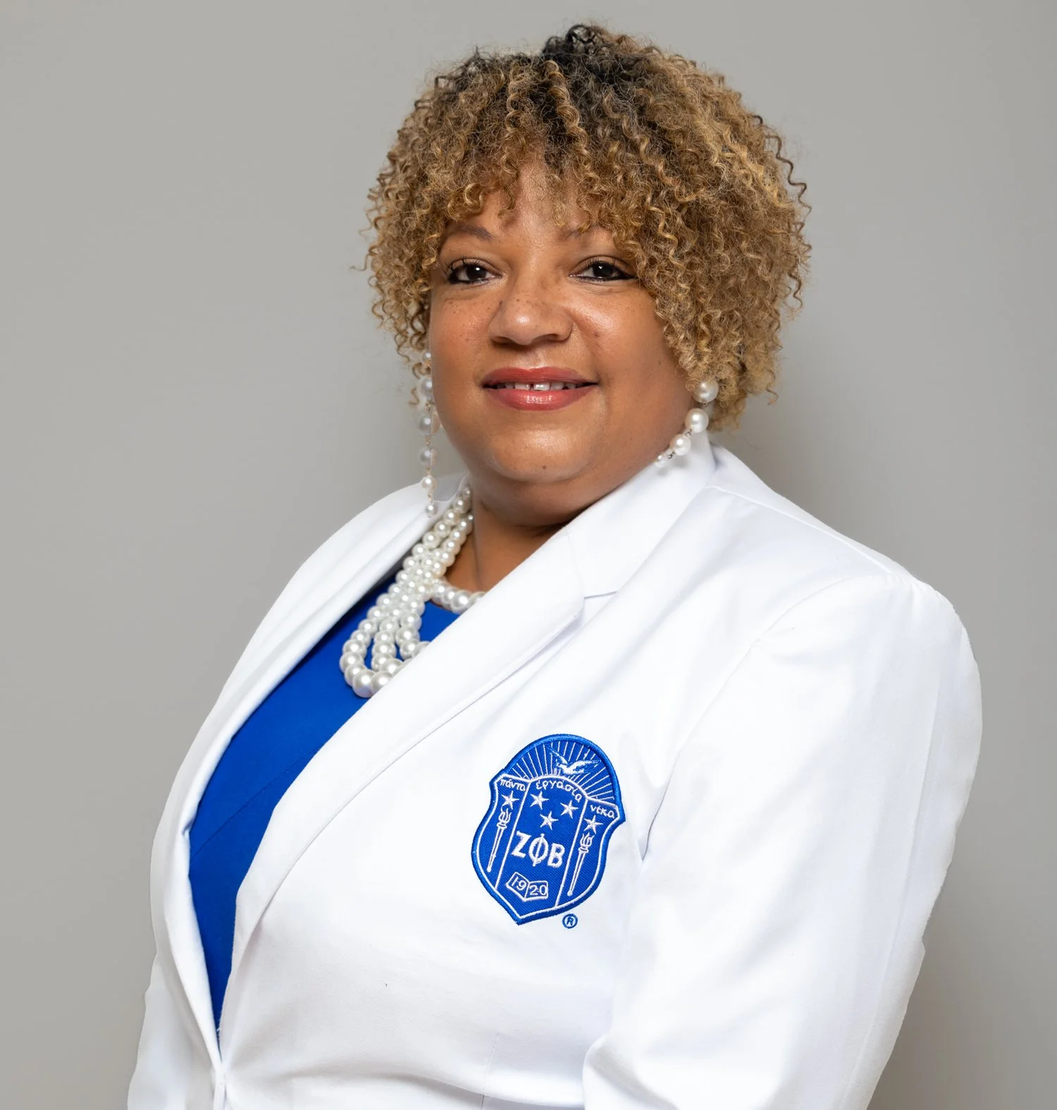 A woman with curly, blonde hair wearing a white blazer with a sorority emblem, pearl earrings, a pearl necklace, and a blue top, posing against a plain gray background.