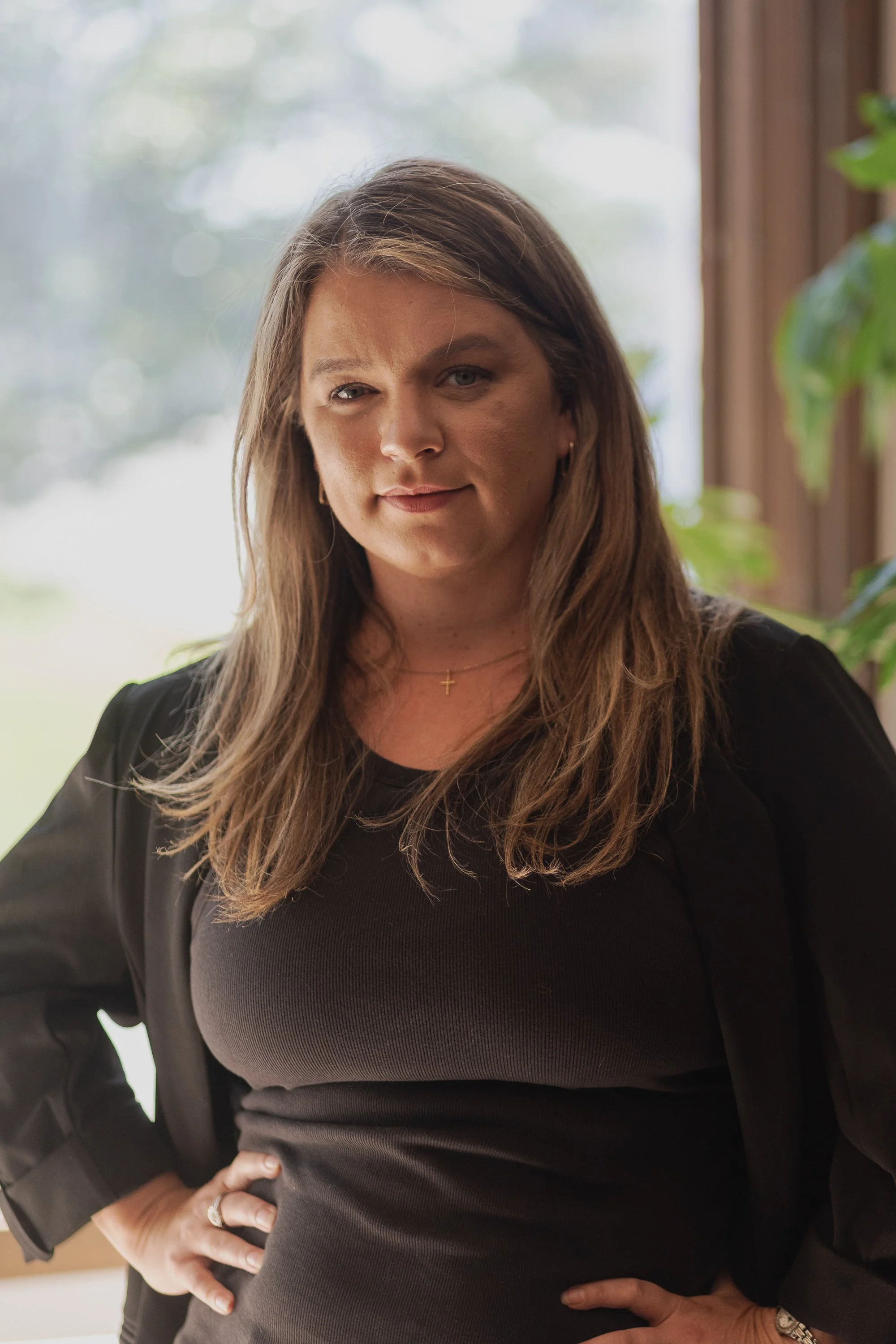 A woman with long brown hair, wearing a black top and gold cross necklace, standing indoors near a window with green plants in the background. She has one hand on her waist and is looking at the camera with a slight smile.