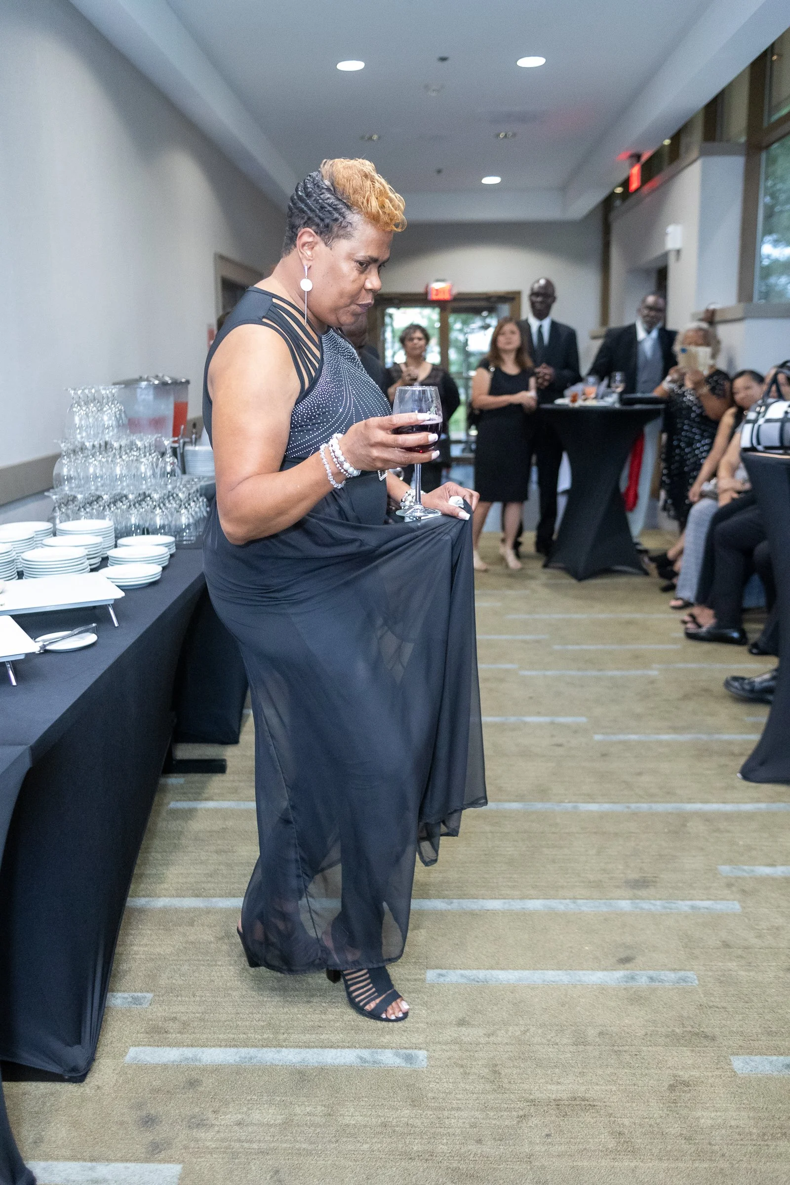 A woman in a black dress holding a glass of red wine, standing in a room with several people in formal attire, near a table with glassware and plates.