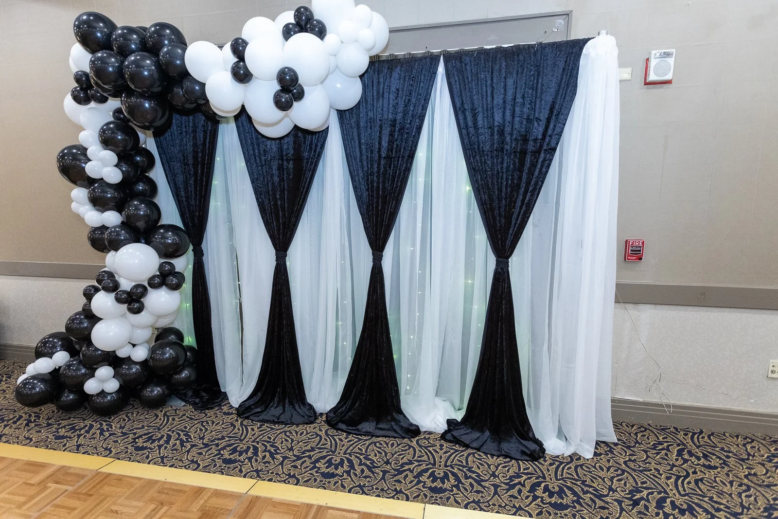 Black and white balloon arrangement with black curtains tied back, white sheer curtains, and fairy lights in the background, set up as an event backdrop.