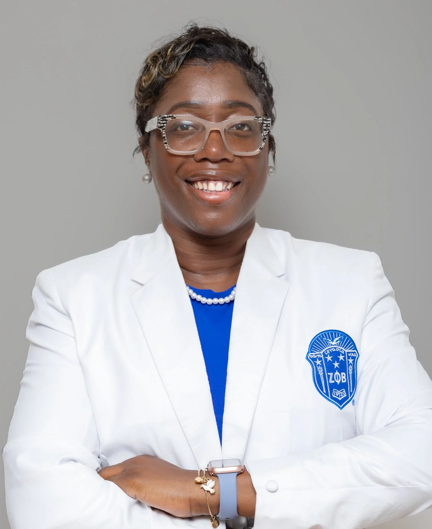 A woman wearing a white blazer with a badge that has Greek letters and a shield. She has glasses, a pearl necklace, and jewelry including a charm bracelet and a smartwatch on her left wrist. She has short wavy hair and is smiling with crossed arms ag