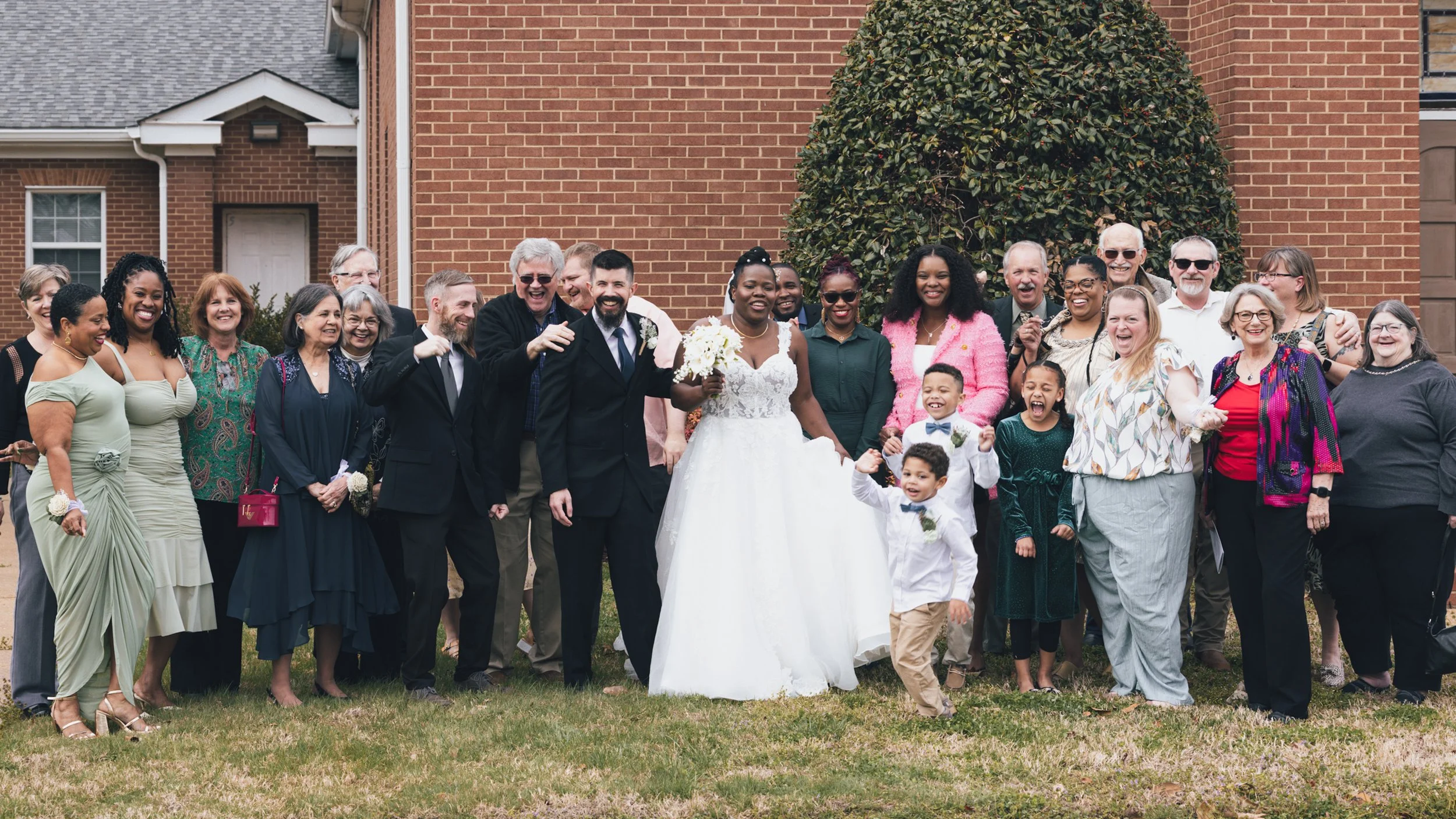 A large group of people, including children, in front of a brick building, celebrating a wedding with the bride in a white gown holding a bouquet and the groom in a suit, all smiling and laughing.
