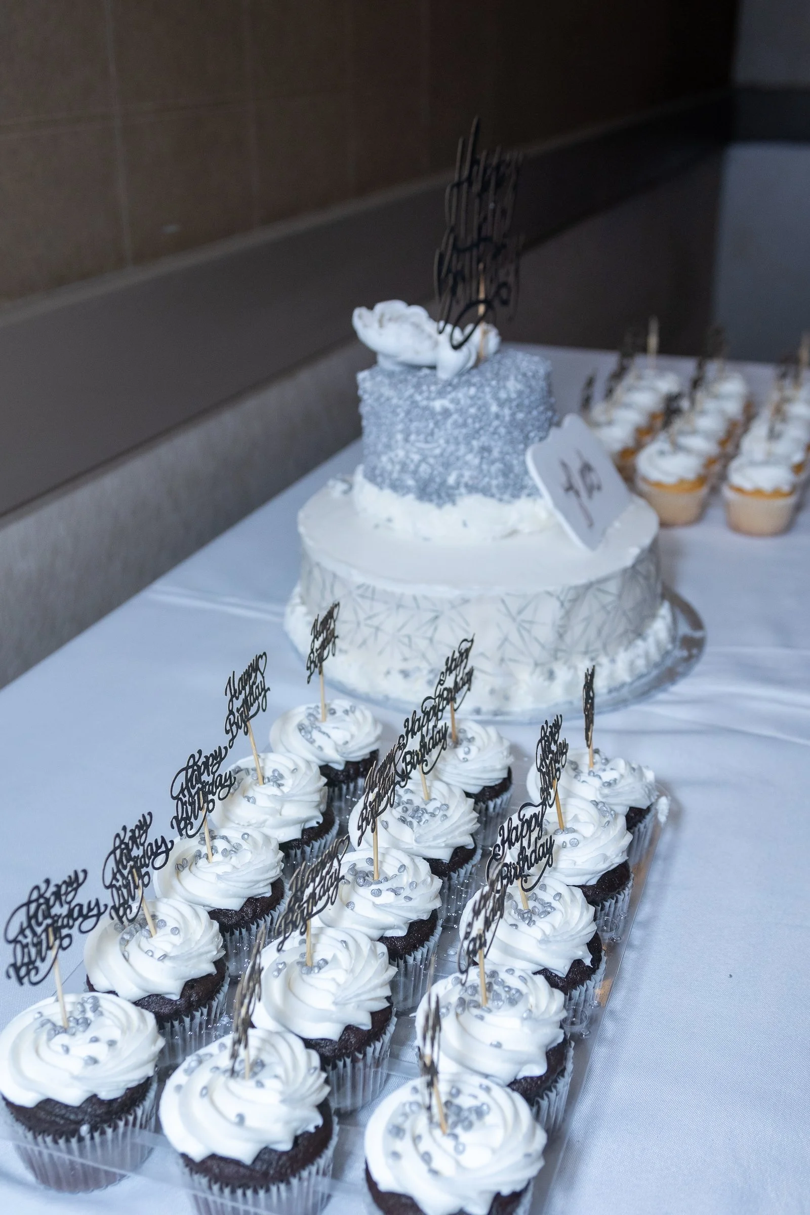 A table with a two-tiered birthday cake decorated with white icing, silver sprinkles, and a swan-shaped decoration on top. Surrounding the cake are cupcakes with white frosting, silver beads, and black 