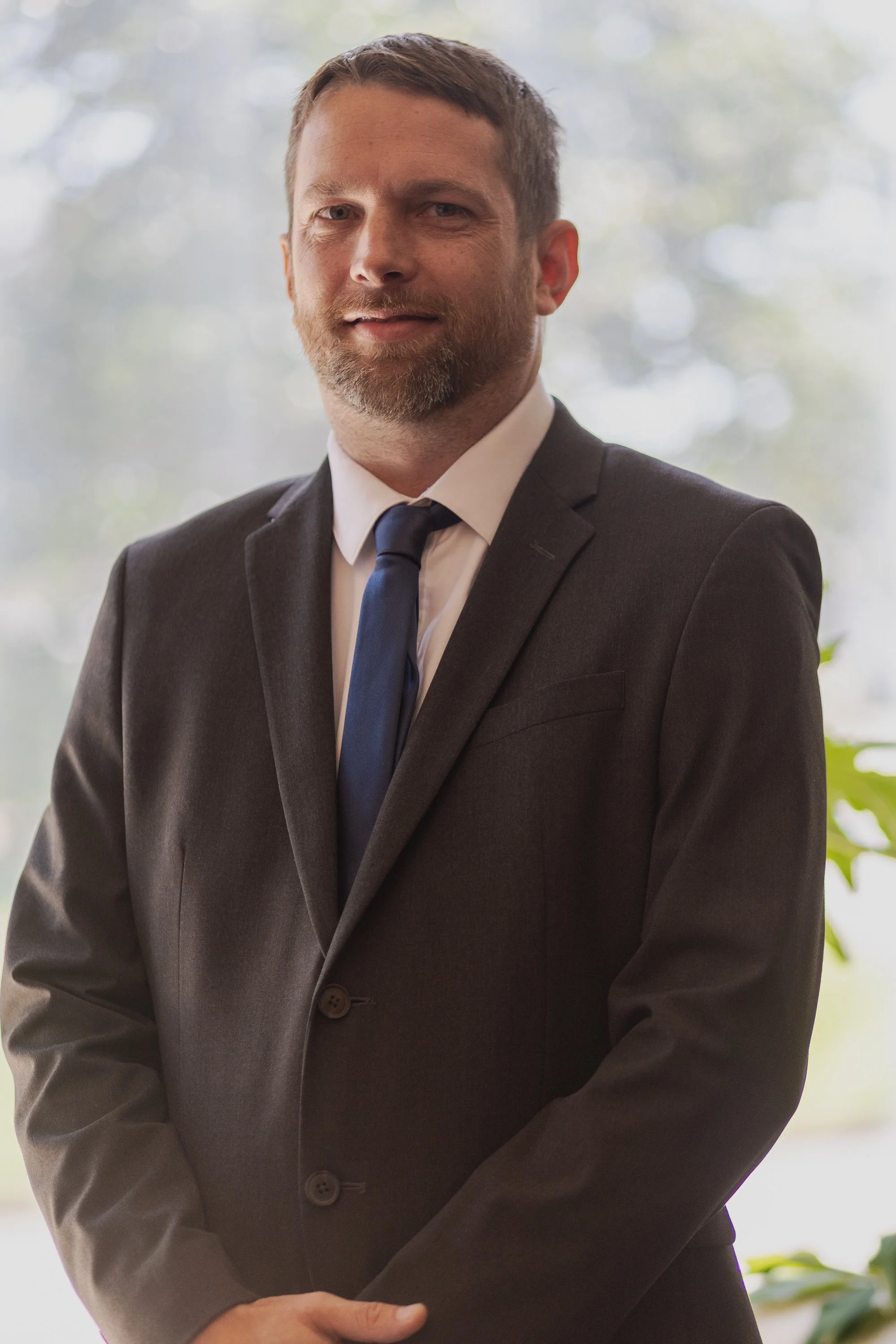 A man in a dark suit, white shirt, and tie standing indoors with a light, blurred background of trees and greenery.