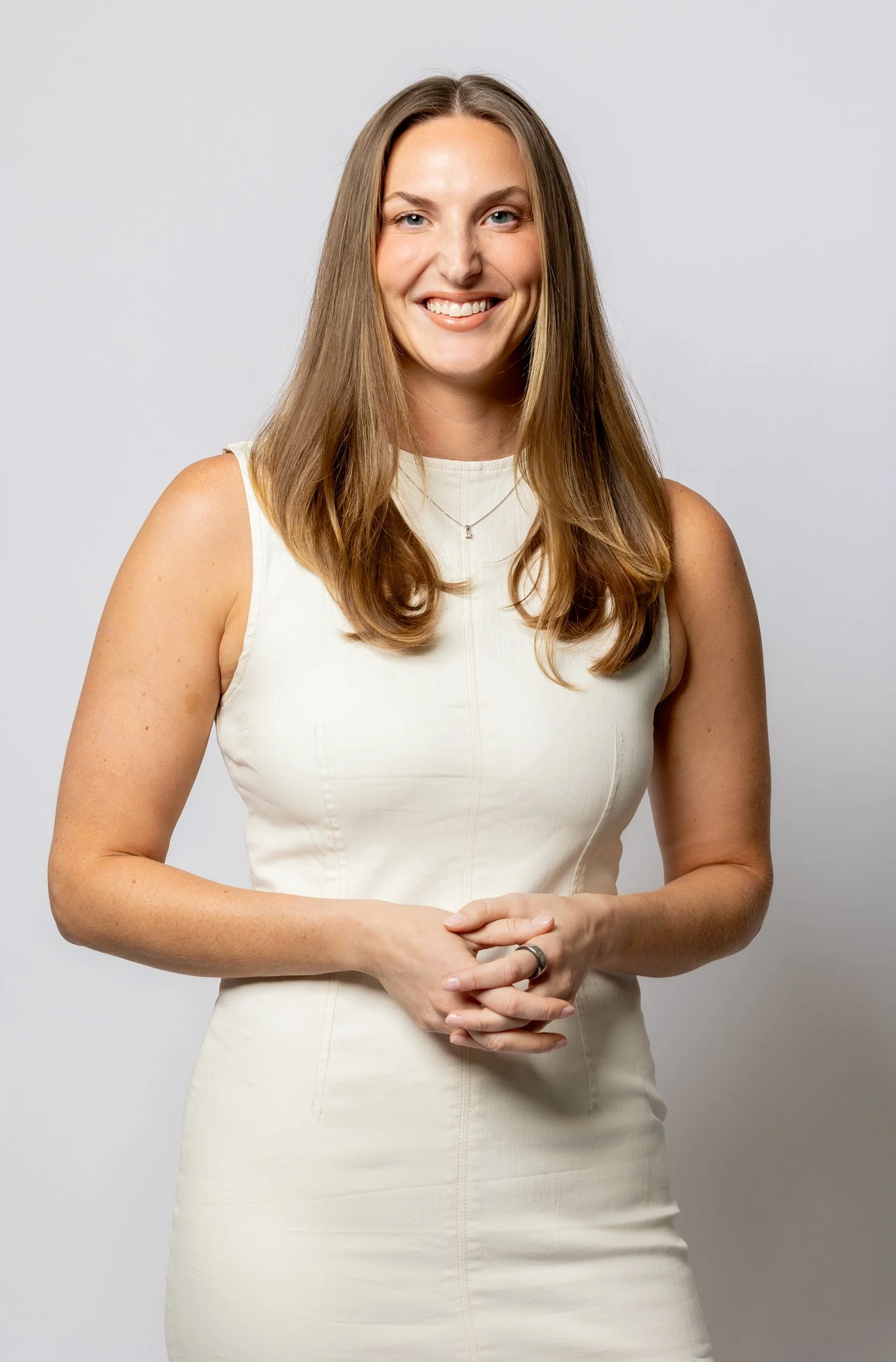 A woman with long brown hair and blue eyes smiling, wearing a white sleeveless dress and a silver necklace, standing against a plain gray background.