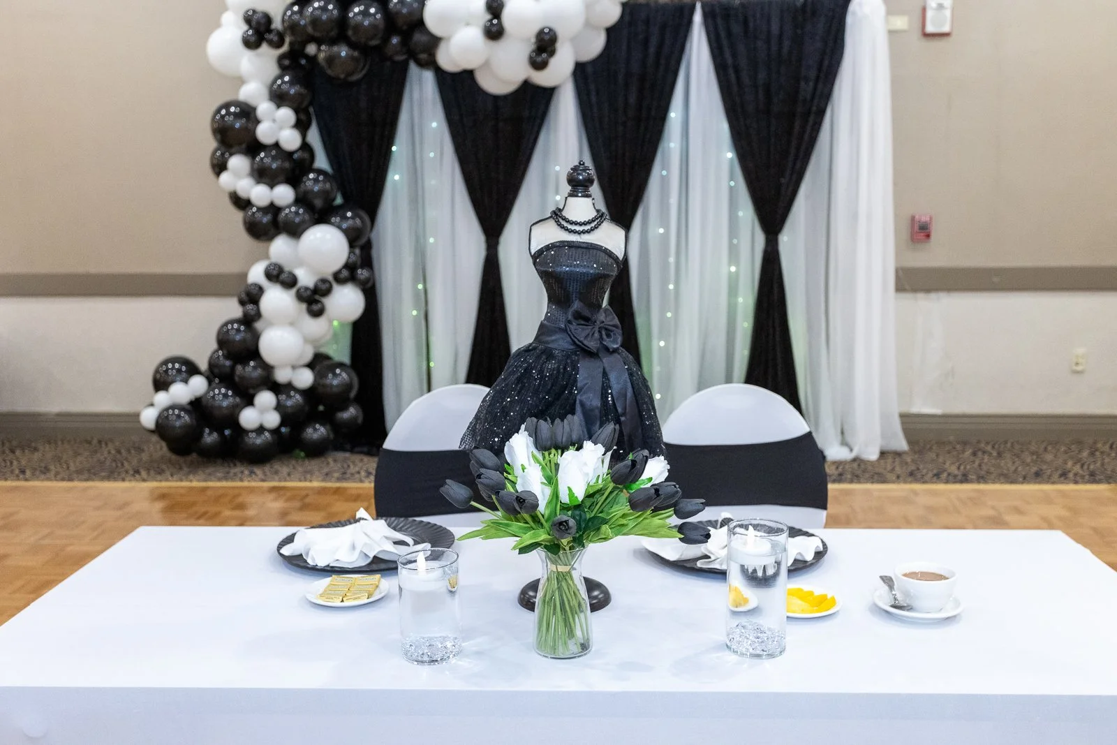 Table setup with a black dress display on a mannequin, a centerpiece of white and black tulips, and balloon decorations in black and white in the background at an event.